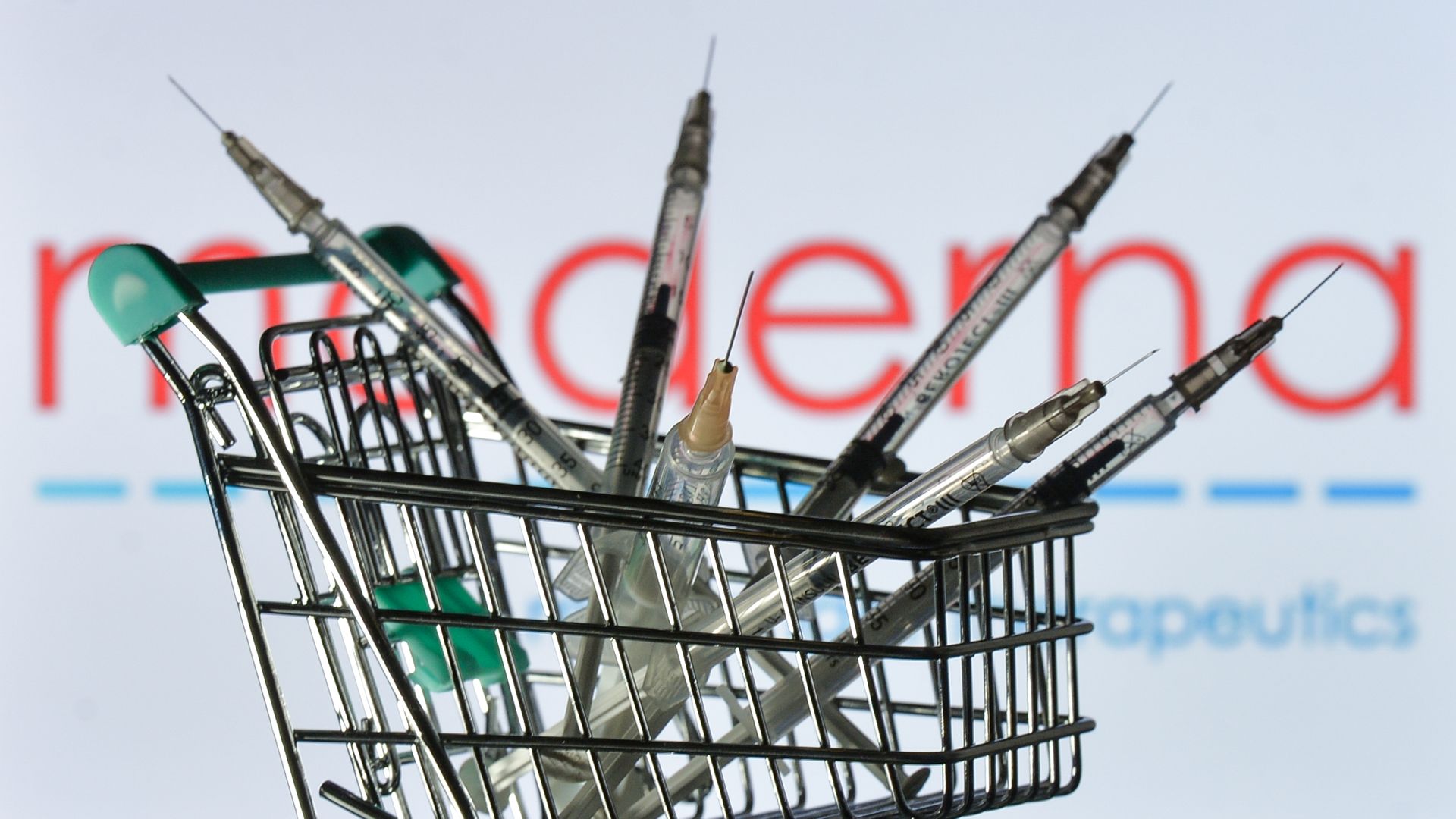 Medical syringes in the mini shopping trolley are seen in front of the Moderna logo displayed on a screen.