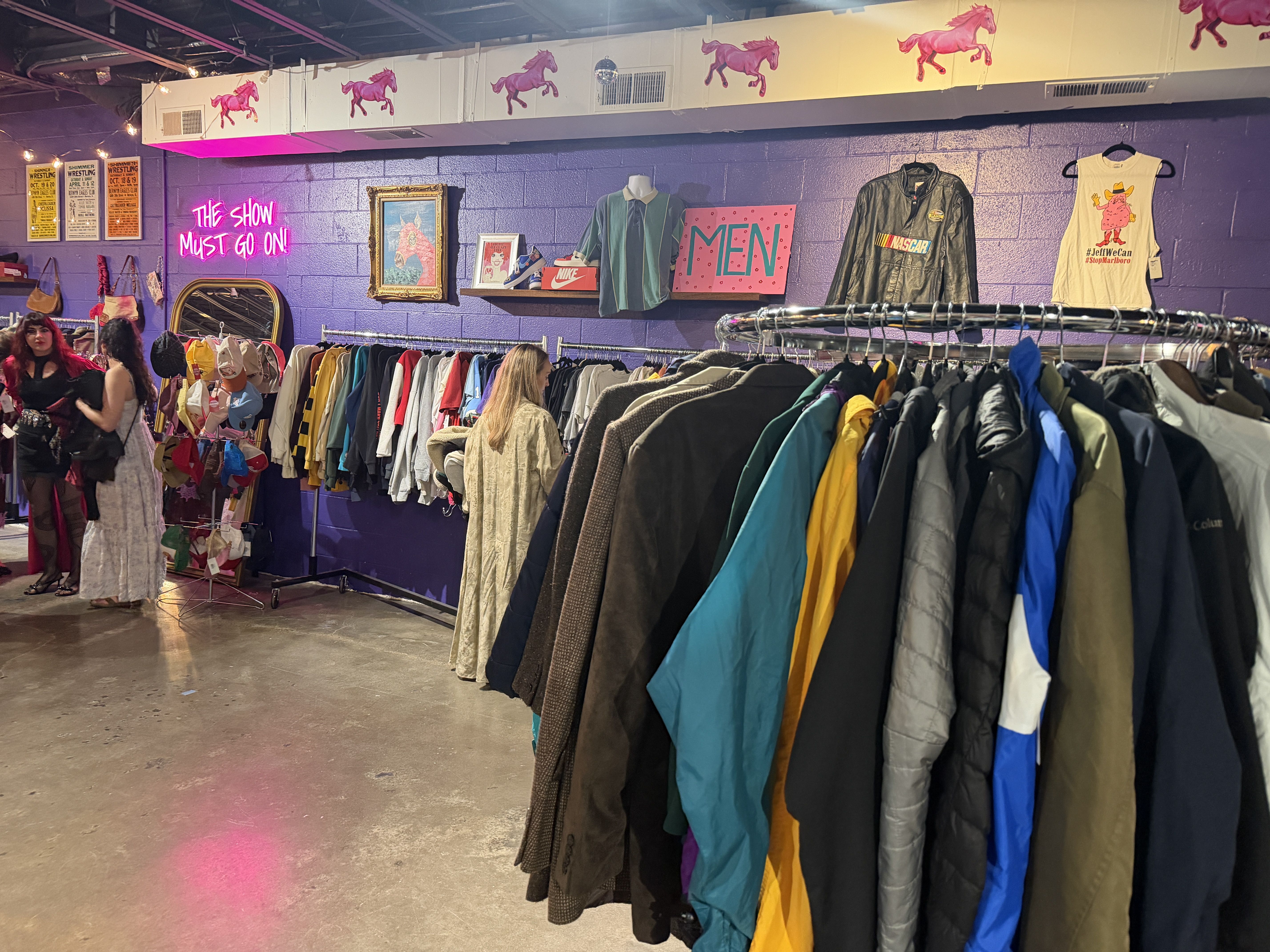 Colorful thrift store interior with a purple wall, racks of coats in blues, greens and yellows; neon pink sign reads THE SHOW MUST GO ON!; shoppers browse near hats and framed art.
