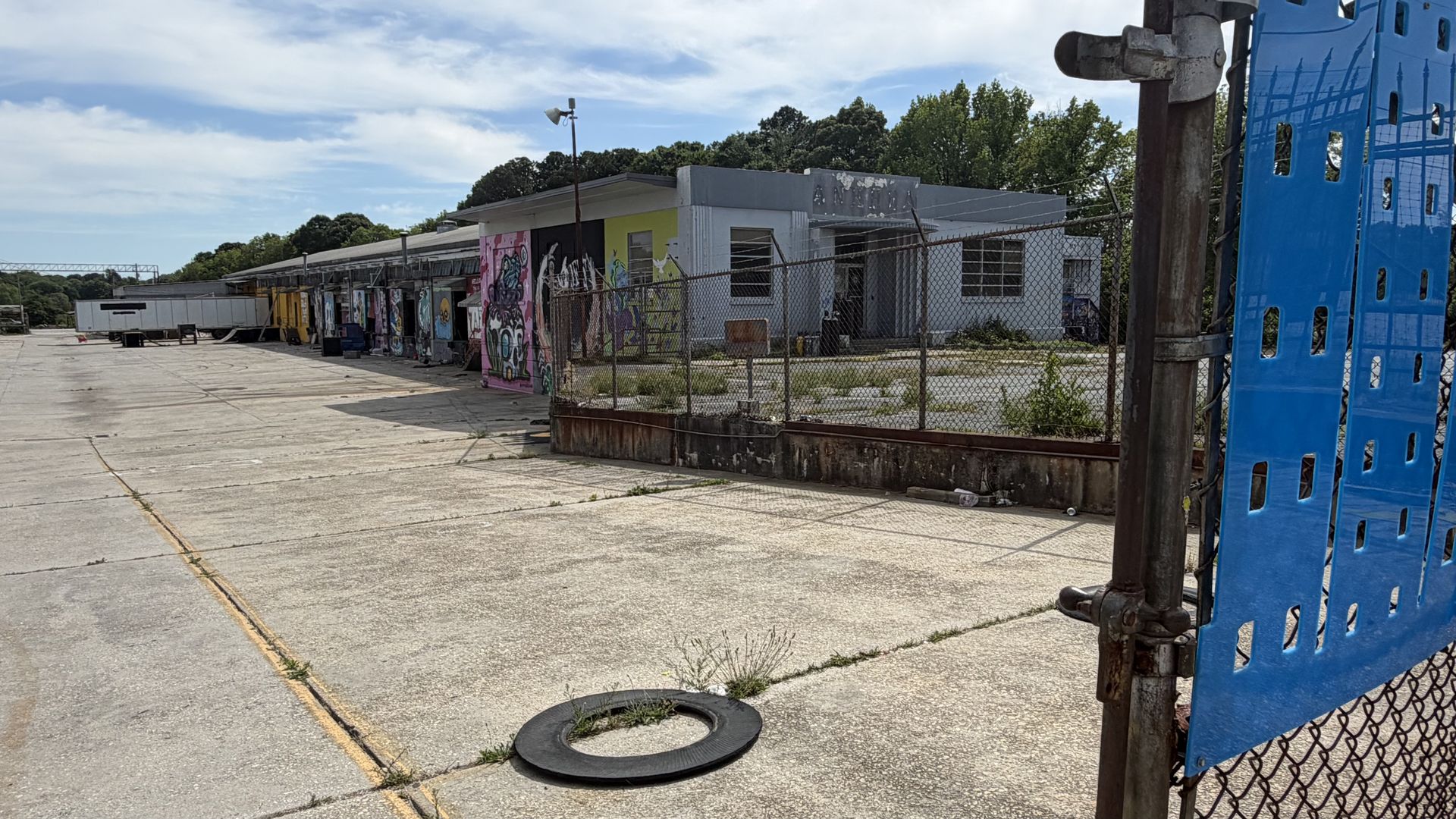 An outdoor loading yard with graffiti-covered trailers and a small grey building behind a chain-link fence. Large blue barriers on the right and scattered tires on cracked concrete under a blue sky.