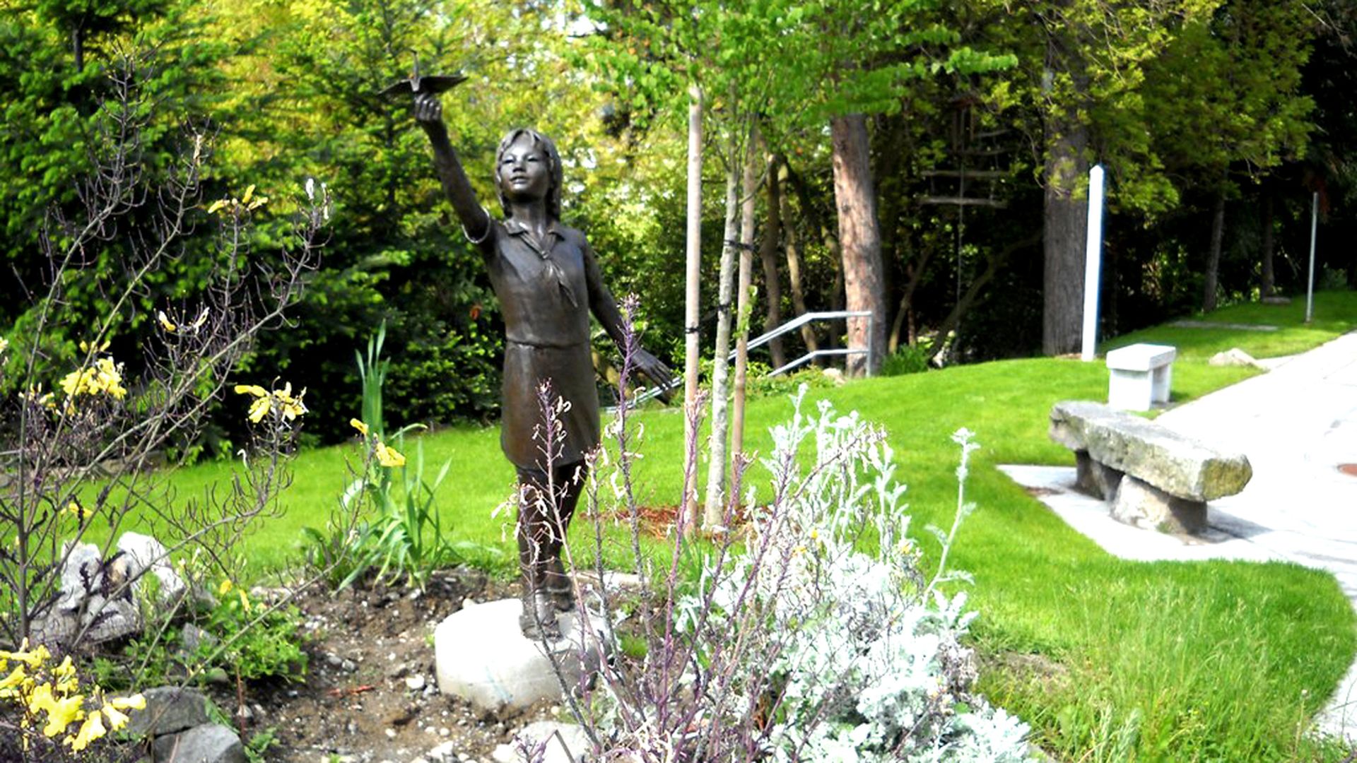 A bronze statue of a girl reaching upward is situated in a green park with a bench and sidewalk at right.
