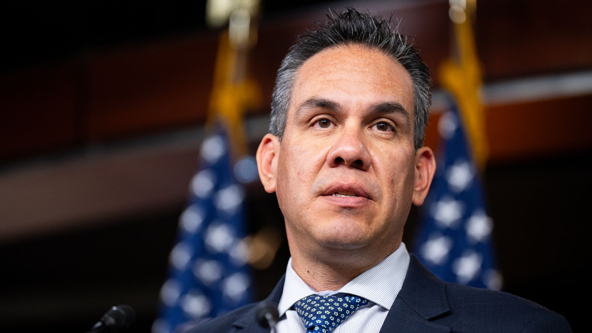 Pete Aguilar, wearing a blue suit and standing in front of American flags.