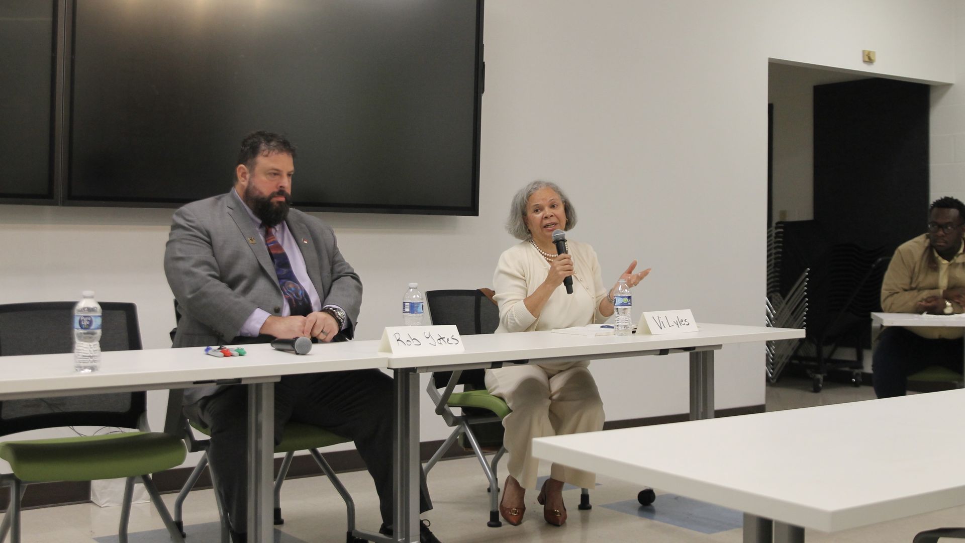 Two panelists seated at a white table in a discussion room, a man in a gray suit and a woman in white holding a microphone, with empty chairs and a third person in the background.