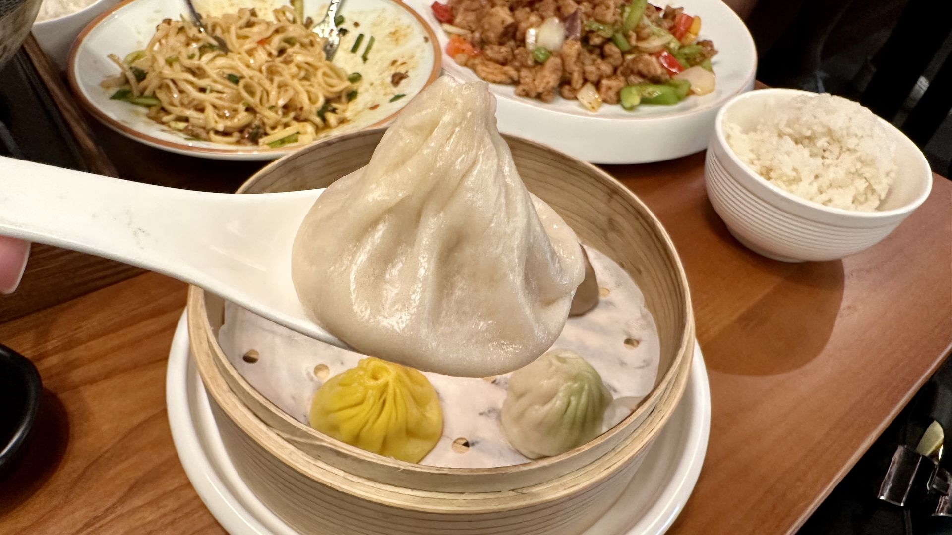 Dim sum meal on a wooden table: a giant white steamed bun in a bamboo steamer, colorful dumplings, noodles and a stir-fry plate, plus a bowl of white rice.