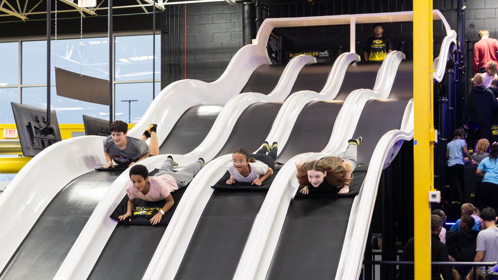 Indoor trampoline park featuring a multi-lane white slide tower; children slide on mats down black lanes as onlookers cluster by yellow pillars and blue railings, with large windows in background.