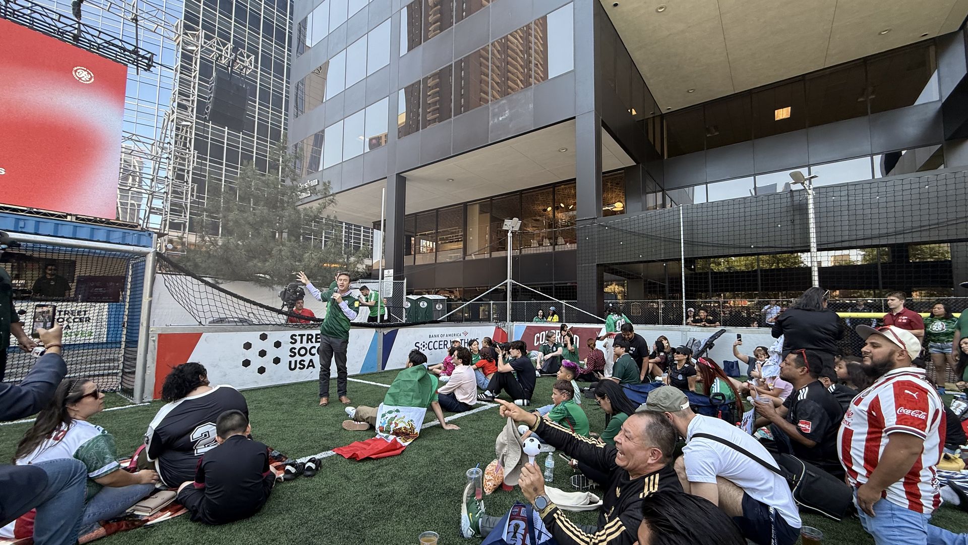 Urban outdoor soccer event on artificial turf, with a large seated crowd behind a barrier. A man in a green shirt speaks into a microphone while cameras film; modern glass skyscrapers reflect in the buildings.