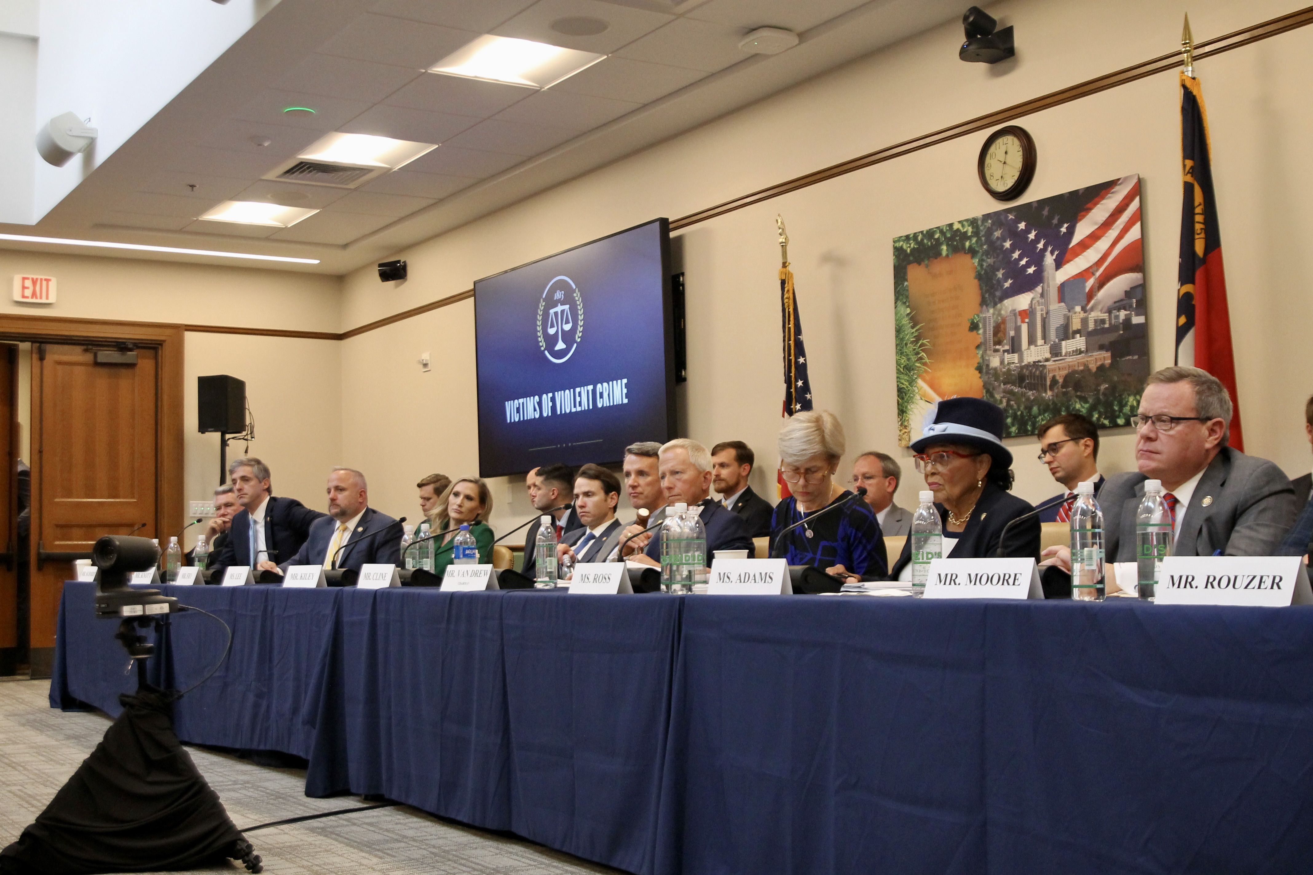 Panel of officials seated at a long table with blue cloth in a conference room, screen behind shows scales and "Victims of Violent Crime," American and state flags visible.