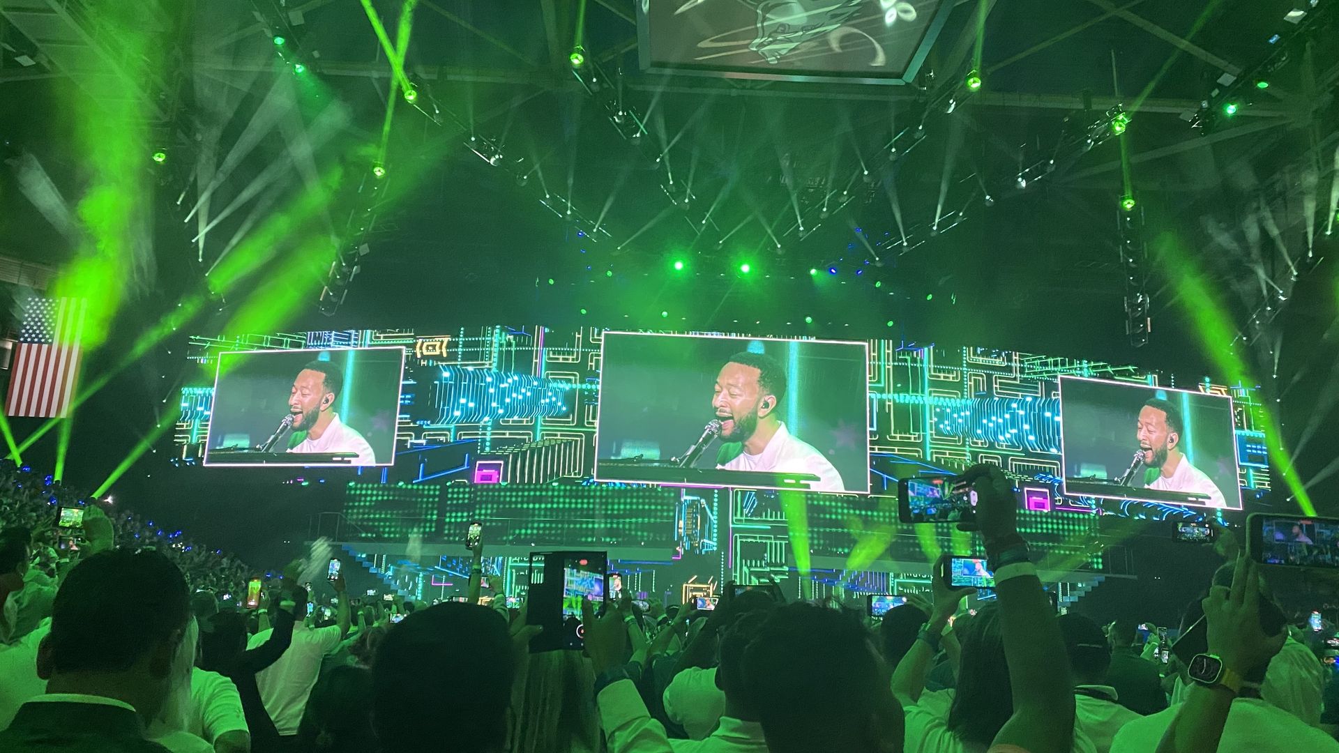 A photo of a crowd watching screens in an arena where John Legend is performing on stage. 