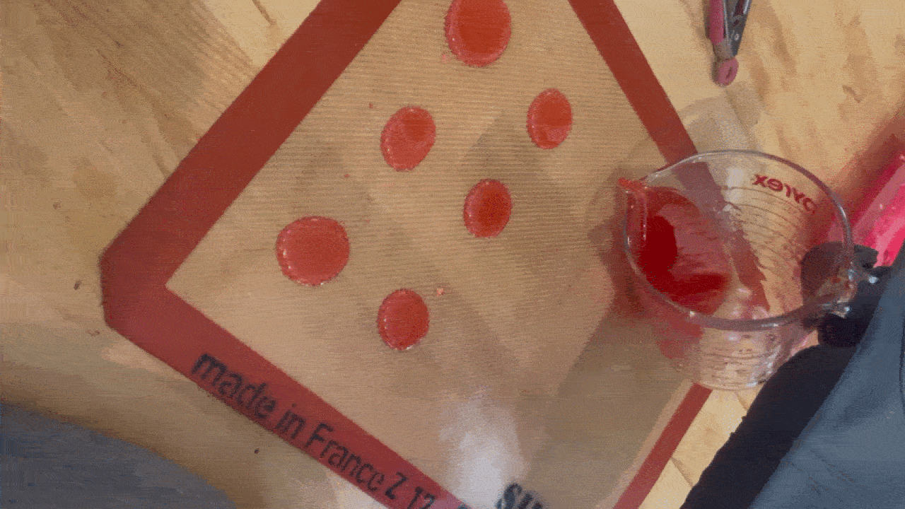 Wooden table with a beige cutting mat showing red polka dots and the words "Made in France" along the edge. A measuring cup with red liquid sits nearby; a pink tool sits in the top-right.