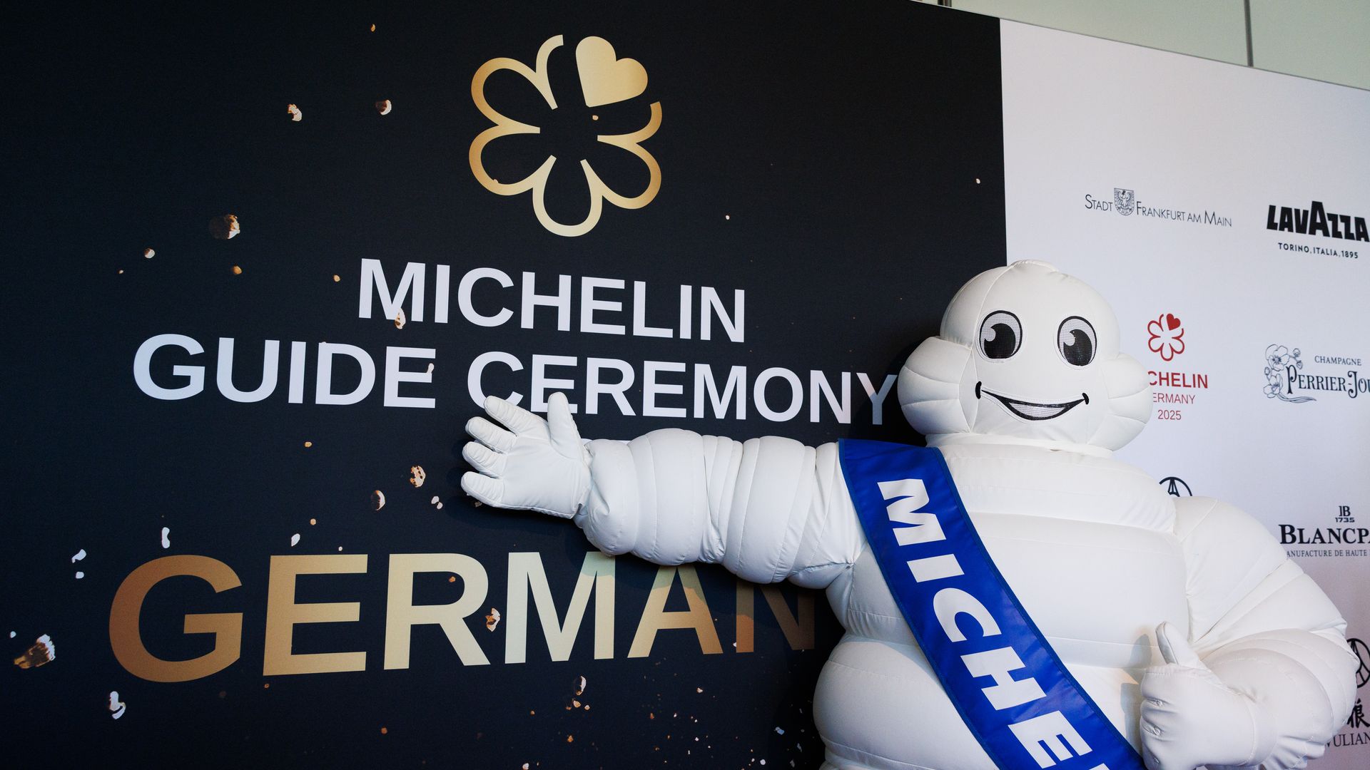 A person dressed up as the Michelin man gestures toward an event sign that says "Michelin Guide Ceremony Germany."