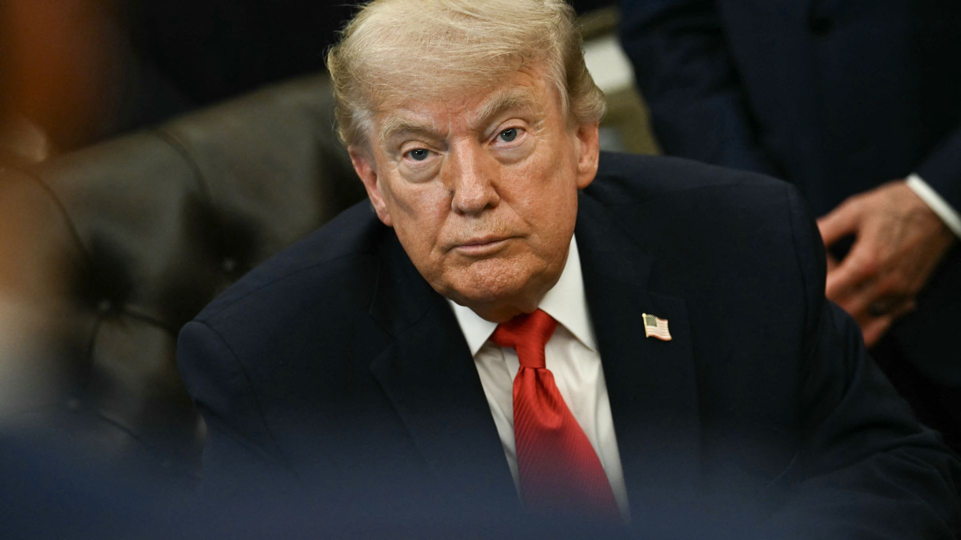 President Donald Trump, wearing a dark suit and red tie, speaks during an Oval Office event at the White House, viewed from across a table with people blurred in the foreground.