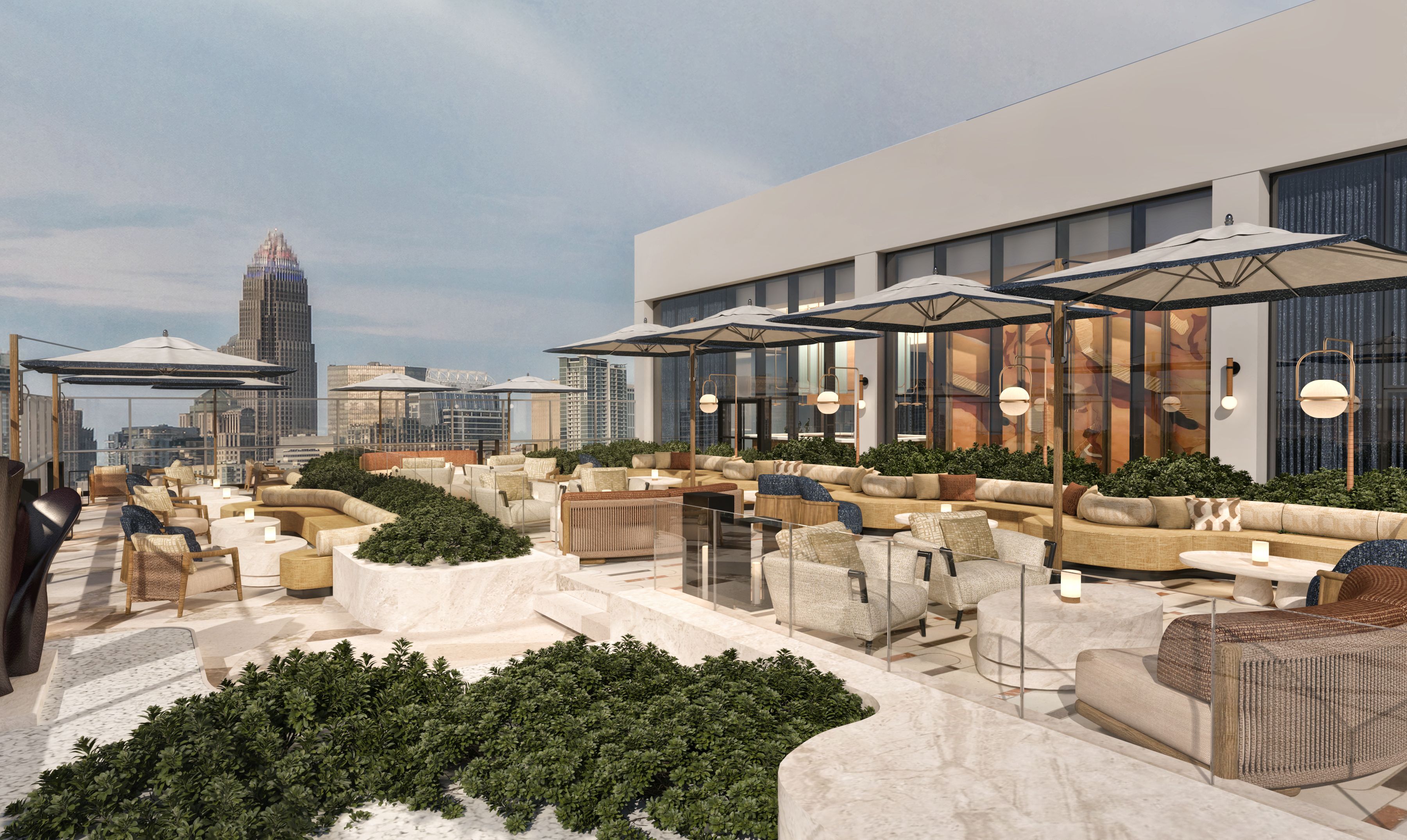 Rooftop lounge terrace with a beige building, cream couches, wicker chairs, and white umbrellas. Glass railing, potted greenery, and round lamps line the seating; city skyline in the distance.