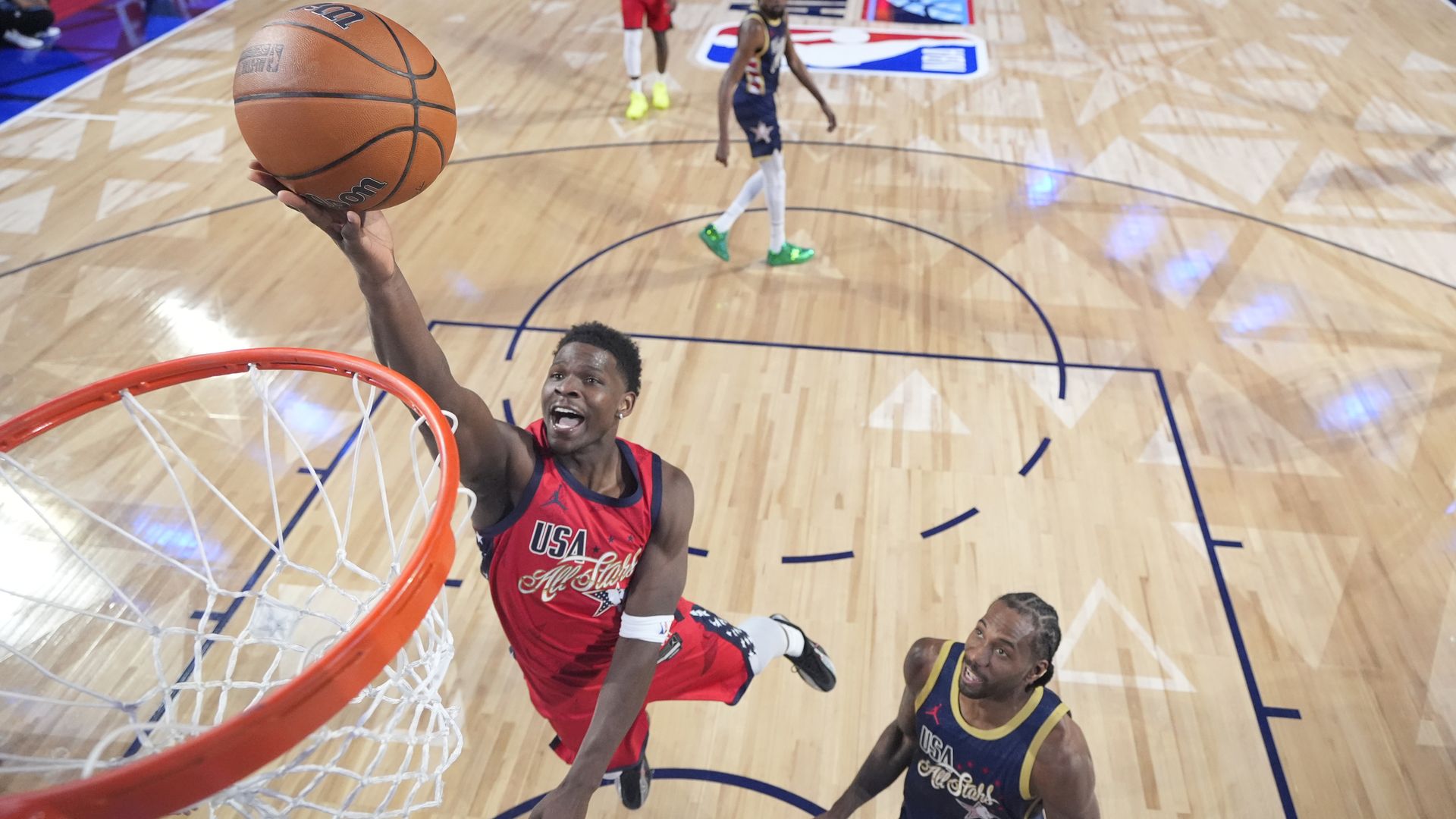 Basketball player Anthony Edwards in red USA All-Star jersey jumps for a layup near the hoop, while a player in blue USA All-Star jersey looks up from below on a polished wood court.
