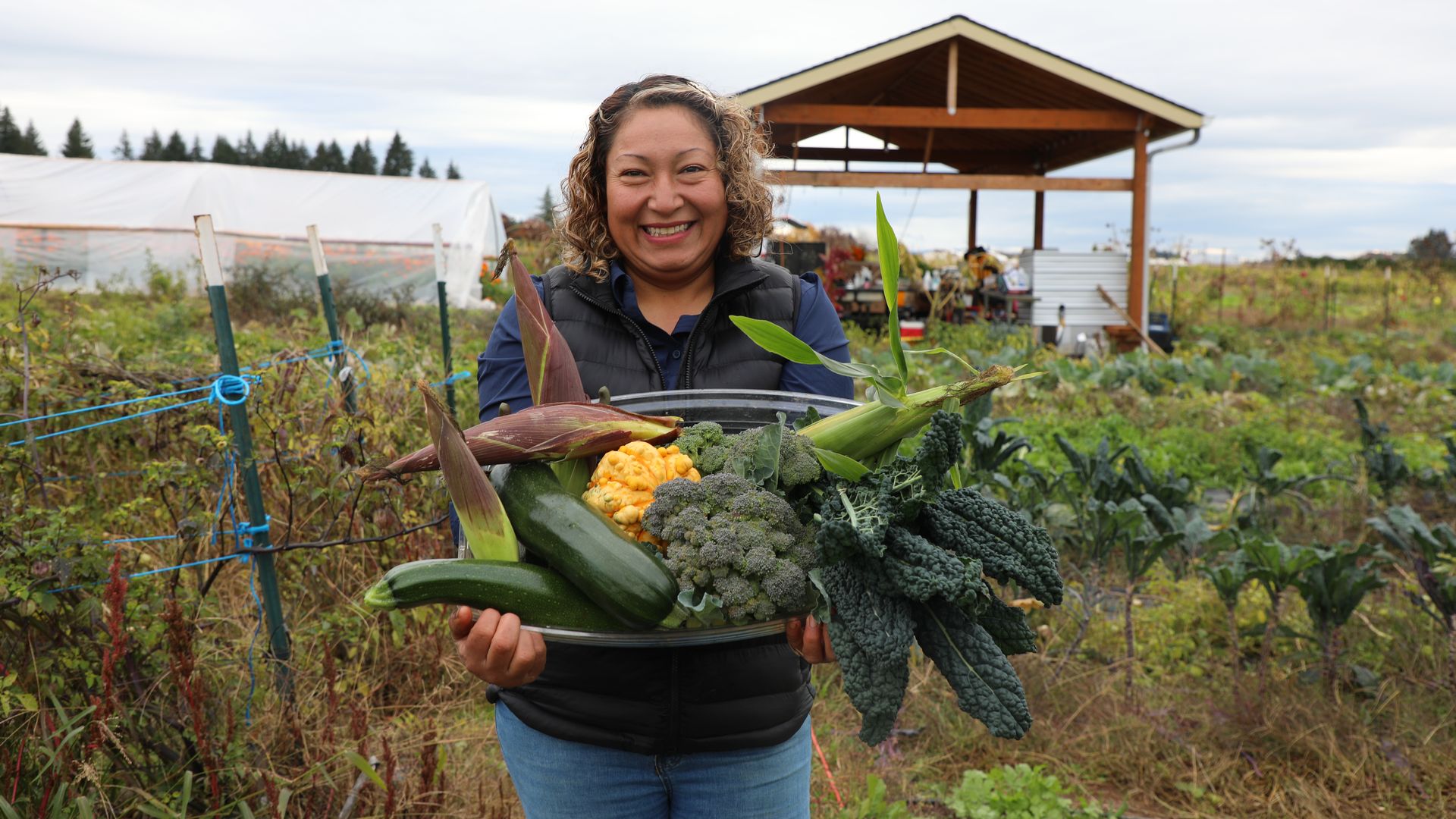 Smiling woman in a navy vest holds a tray of fresh vegetables (corn, broccoli, kale, squash) in a rural garden with a wooden shelter and greenhouse in the background.