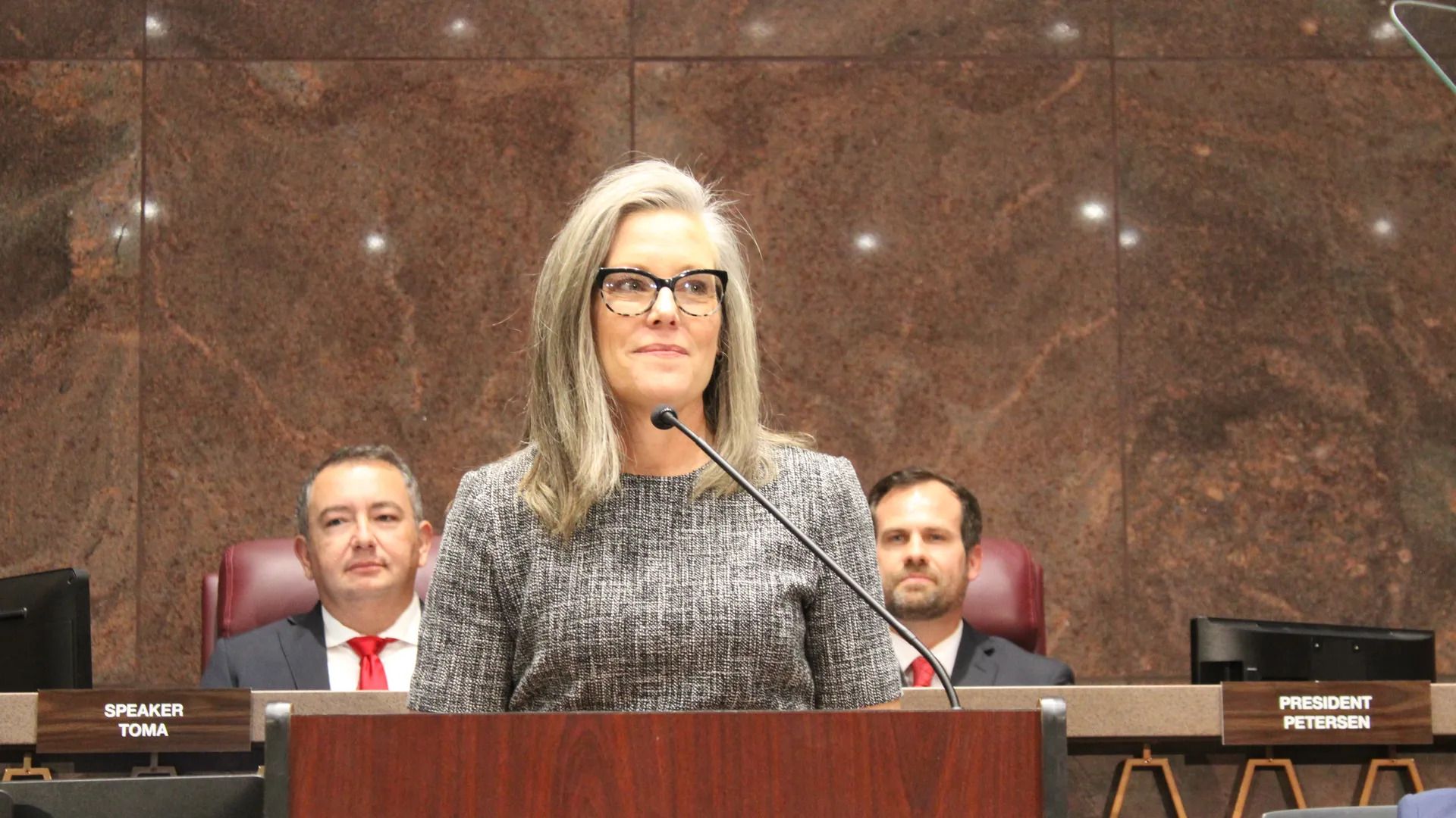 A woman stands in front of a microphone at a lectern with two men sitting behind her. 