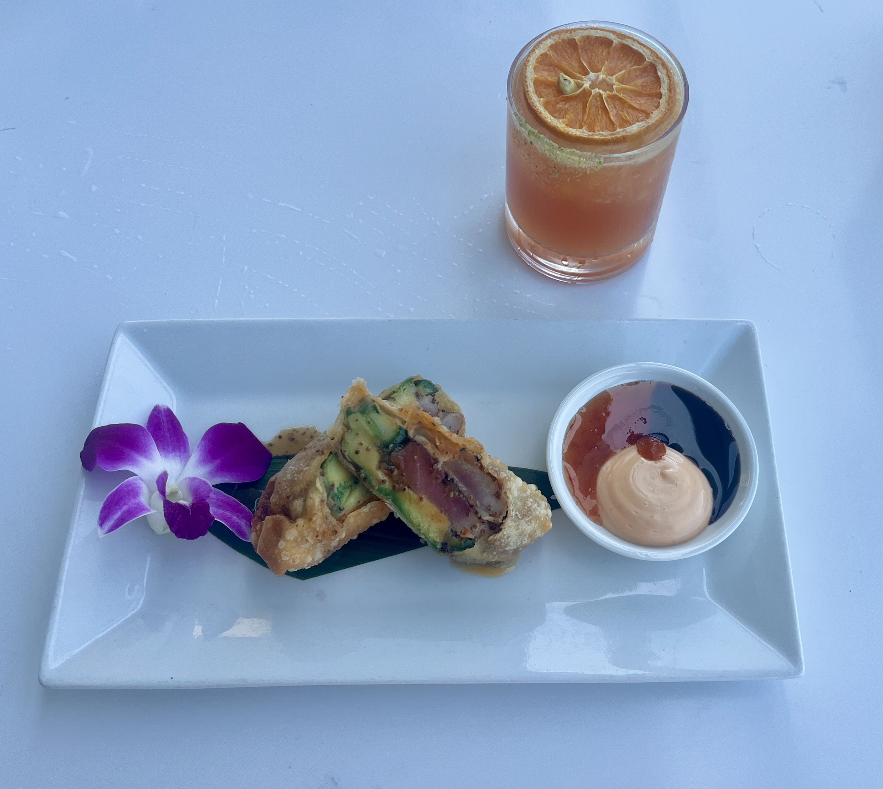 White rectangular plate with two crispy fried spring rolls filled with avocado and tuna, purple orchid flower, and a small bowl of three dipping sauces; a pink cocktail with a dried orange slice on top is beside the plate.