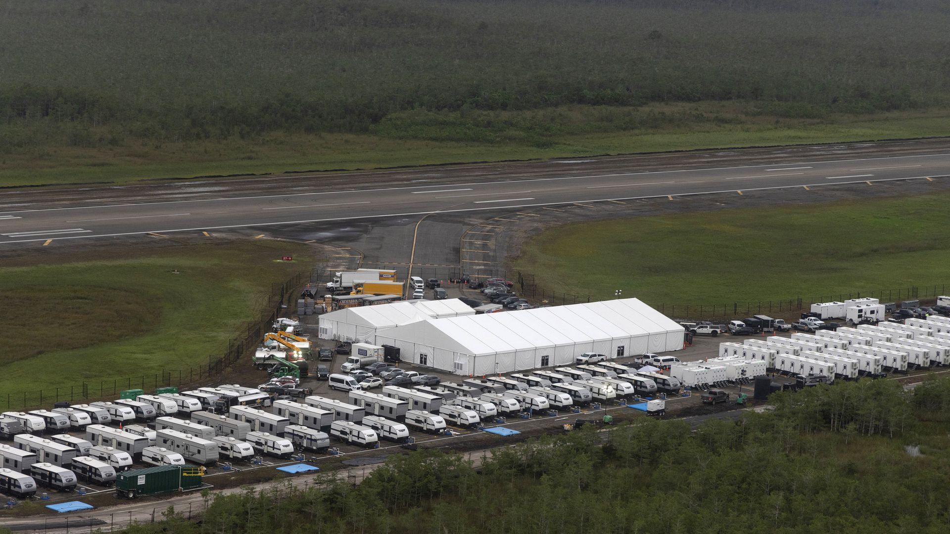 Aerial view of a large temporary camp with many white trailers and two large white tents next to a runway, surrounded by green fields and forested areas.