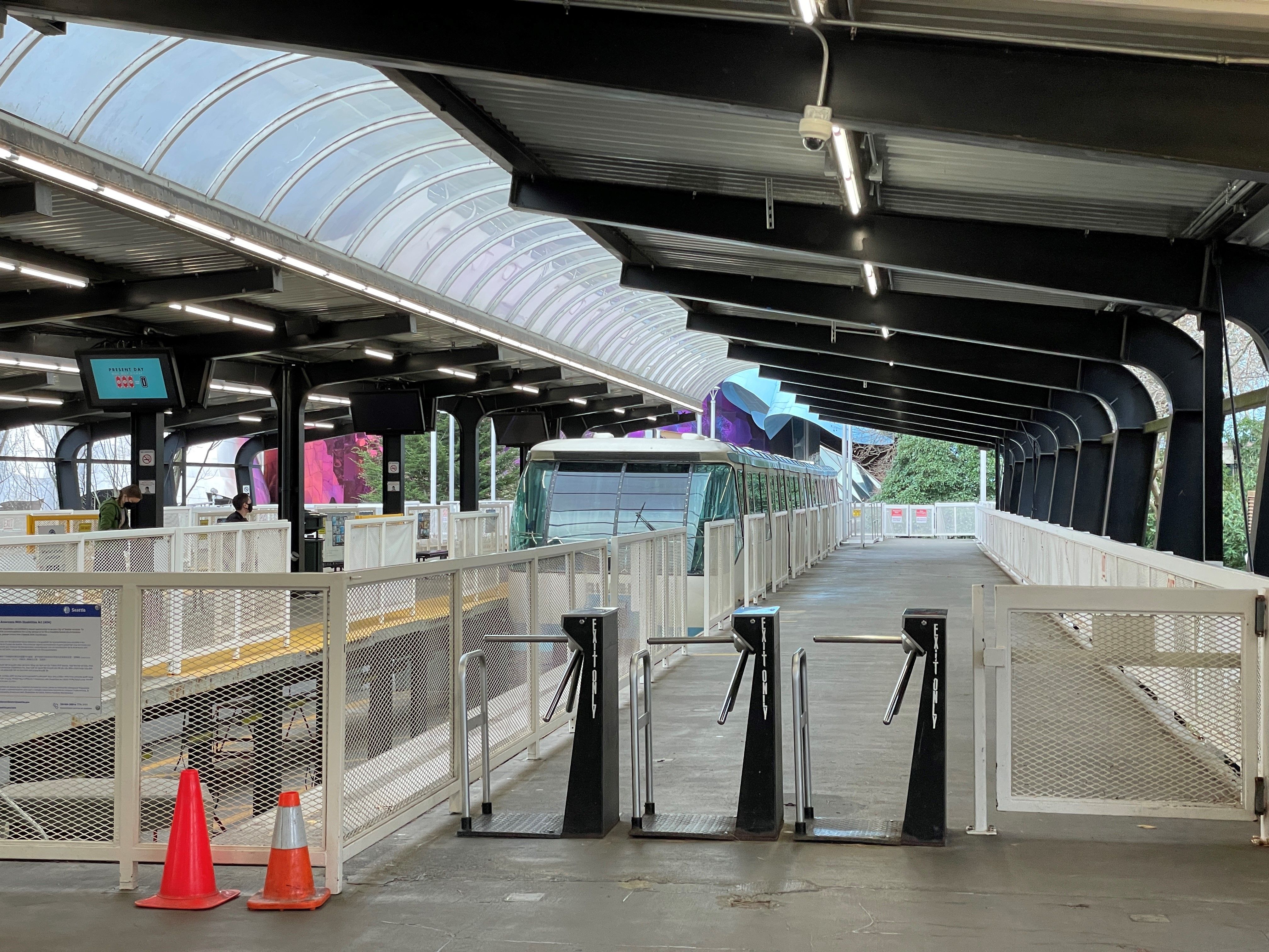 Empty train station platform with white metal fences, three turnstiles labeled "Exit Only," two orange traffic cones, and a stationary train under a transparent arched roof.