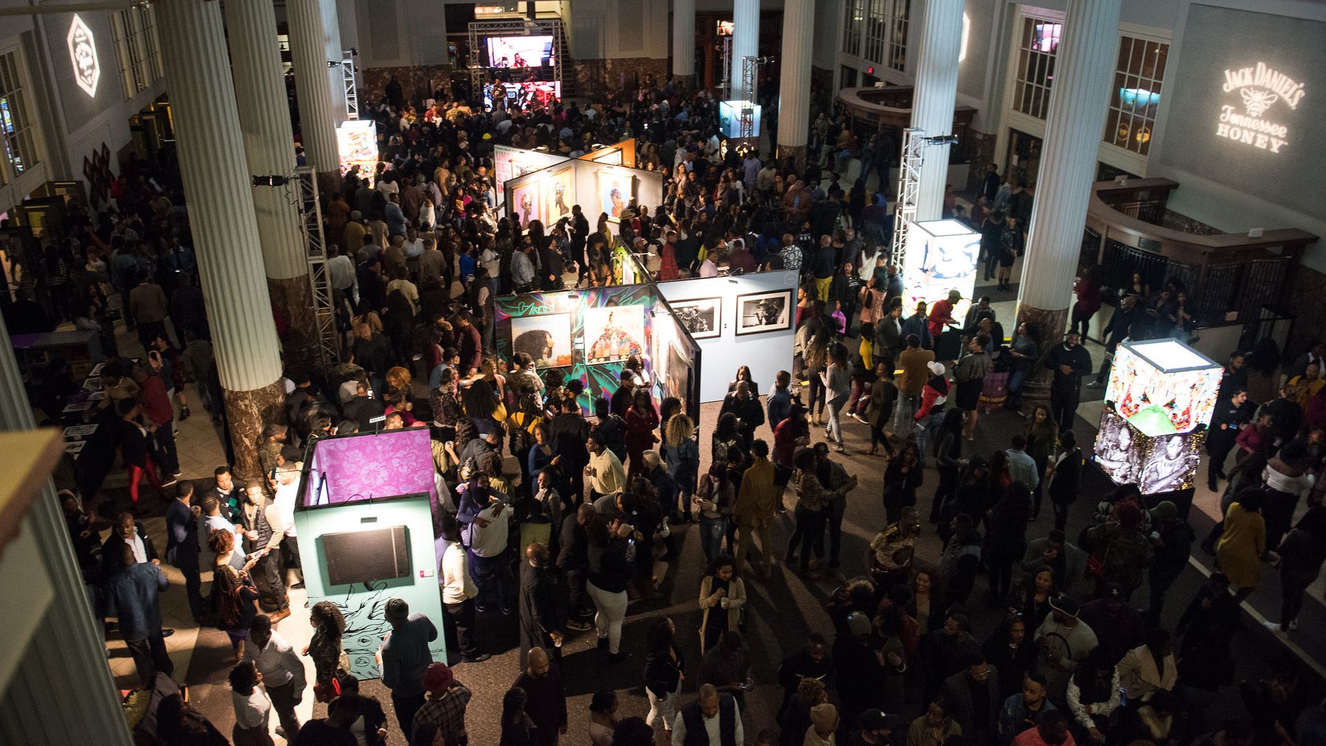 Art, Beats + Lyrics coming to charlotte Crowded indoor art exhibition in a large hall with tall white columns, illuminated art displays, and a projected Jack Daniel's Tennessee Honey logo.