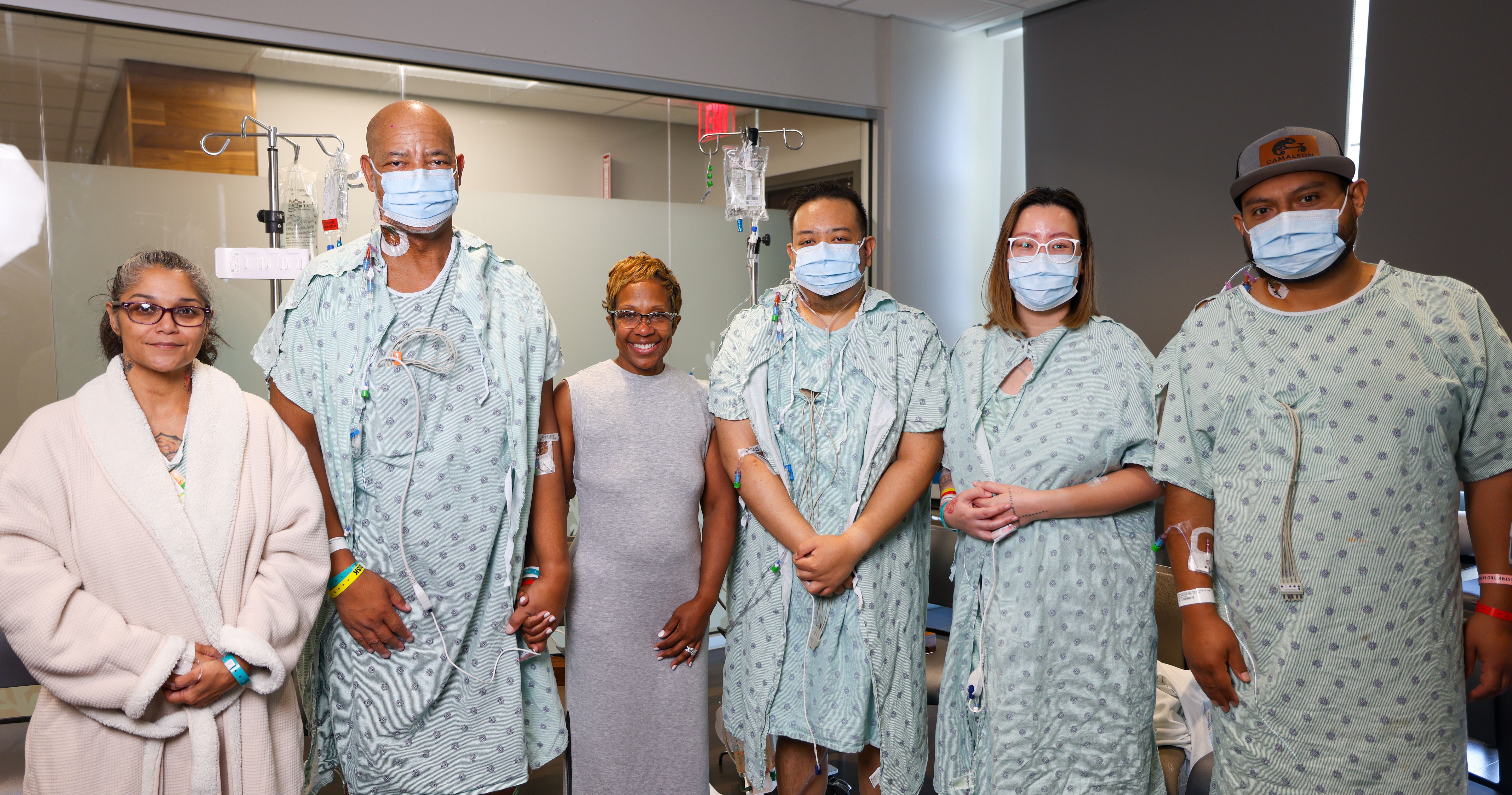 Group of seven people in a hospital ward posing for a photo; most wear light blue patient gowns and masks, one woman wears a gray sleeveless top; IV poles and equipment visible in background.