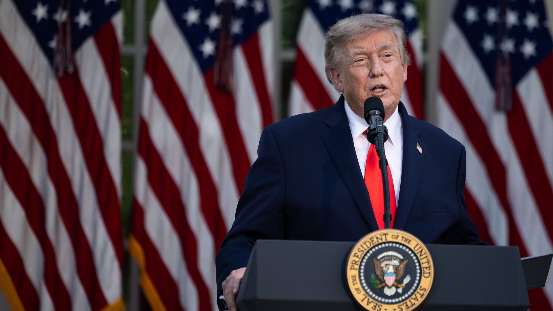 President Trump in a dark suit and red tie speaks at a podium with the presidential seal, microphone in front, American flags in the background.