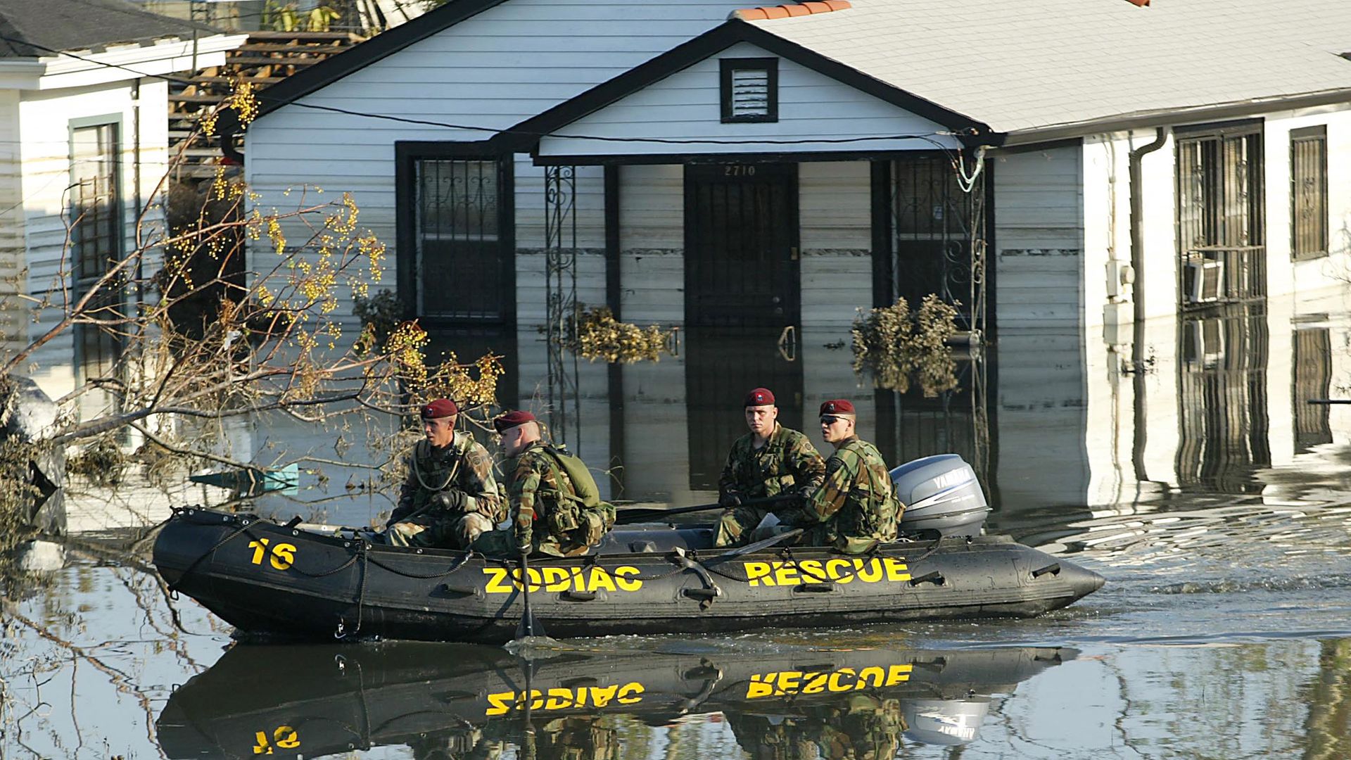 Image shows soldiers in an inflatable raft in floodwaters. A flooded house is in the background.