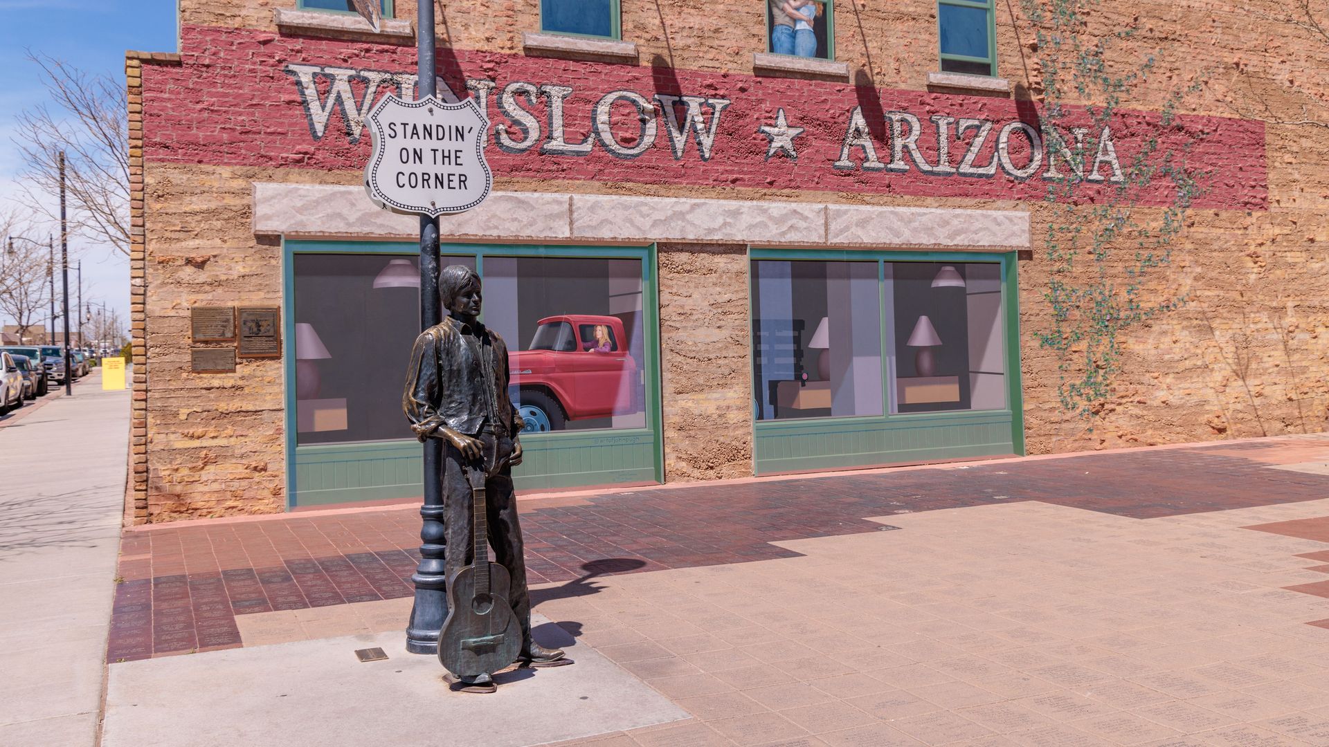 Bronze guitarist statue on a lamppost in front of a brick building with a red mural reading "SLOW ARIZONA." A sign on the pole says "STANDIN ON THE CORNER." Green-framed storefront windows, ivy, and a red vintage truck inside.
