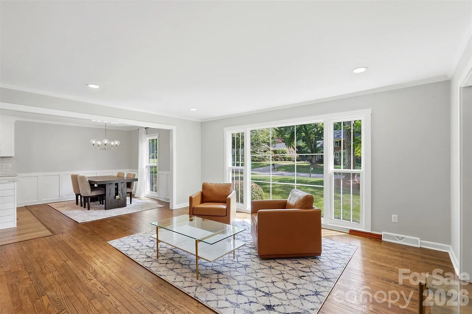 Bright, open living room with gray walls, white trim, and large front windows. Two tan leather chairs flank a glass coffee table on a patterned rug; dining area with a dark wood table in the back.