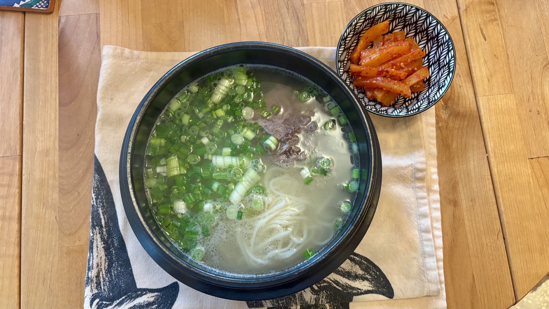 Bowl of Korean soup with noodles, beef, and chopped green onions, next to a small bowl of red kimchi on a wooden table with a white cloth featuring a black graphic.