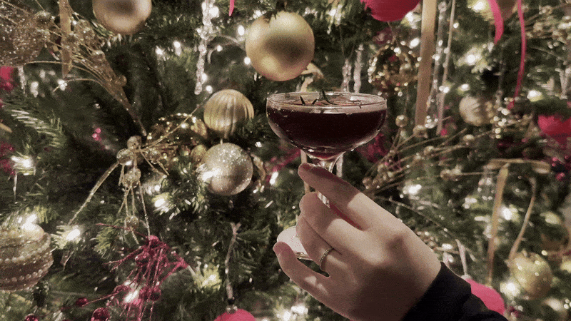 Image shows a woman's hand toasting with a cocktail in front of a Christmas tree.