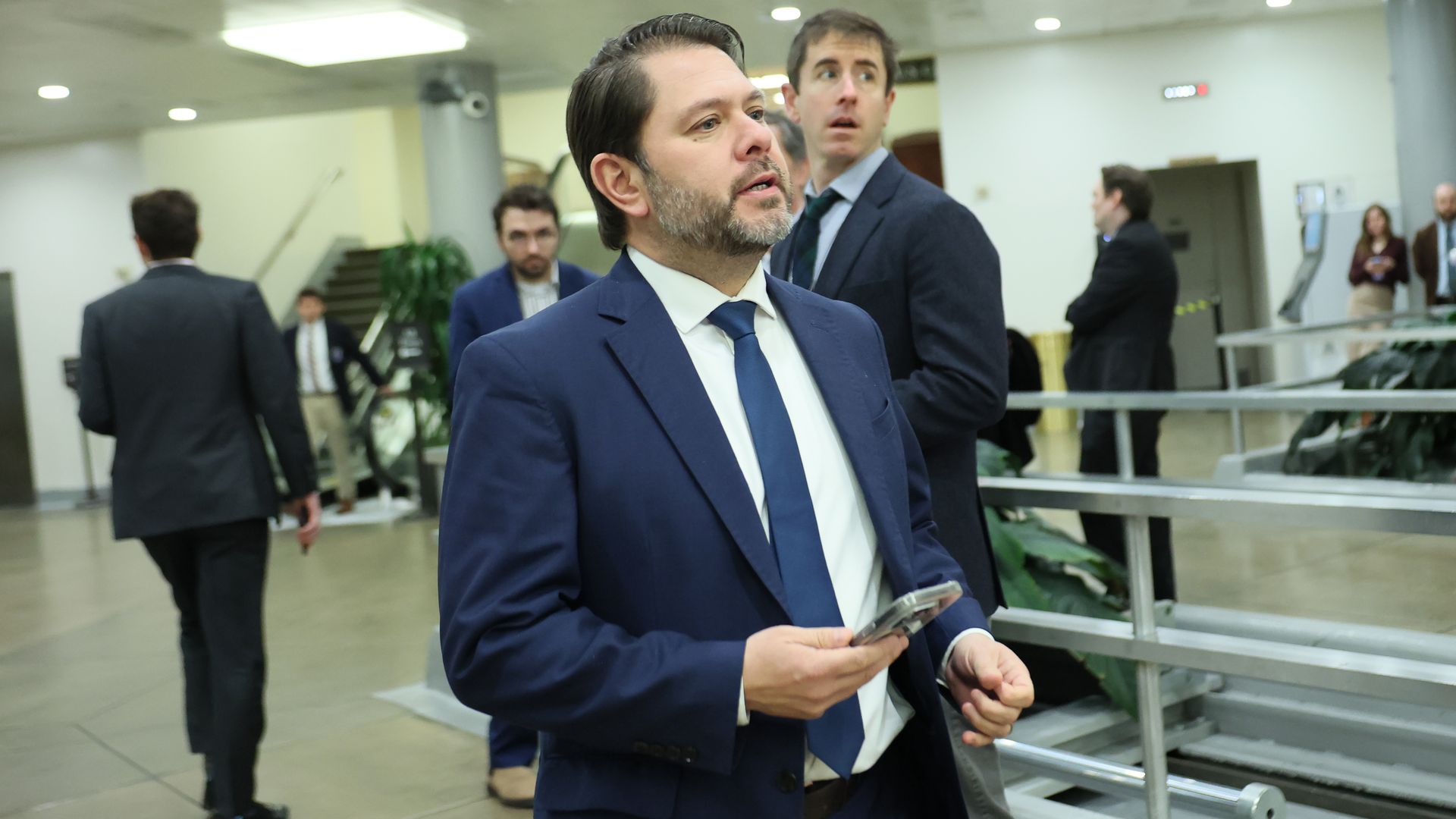 A man in a navy suit and blue tie walks through a modern office lobby, holding a smartphone; behind him, several suited colleagues stand near a railing and stairs.