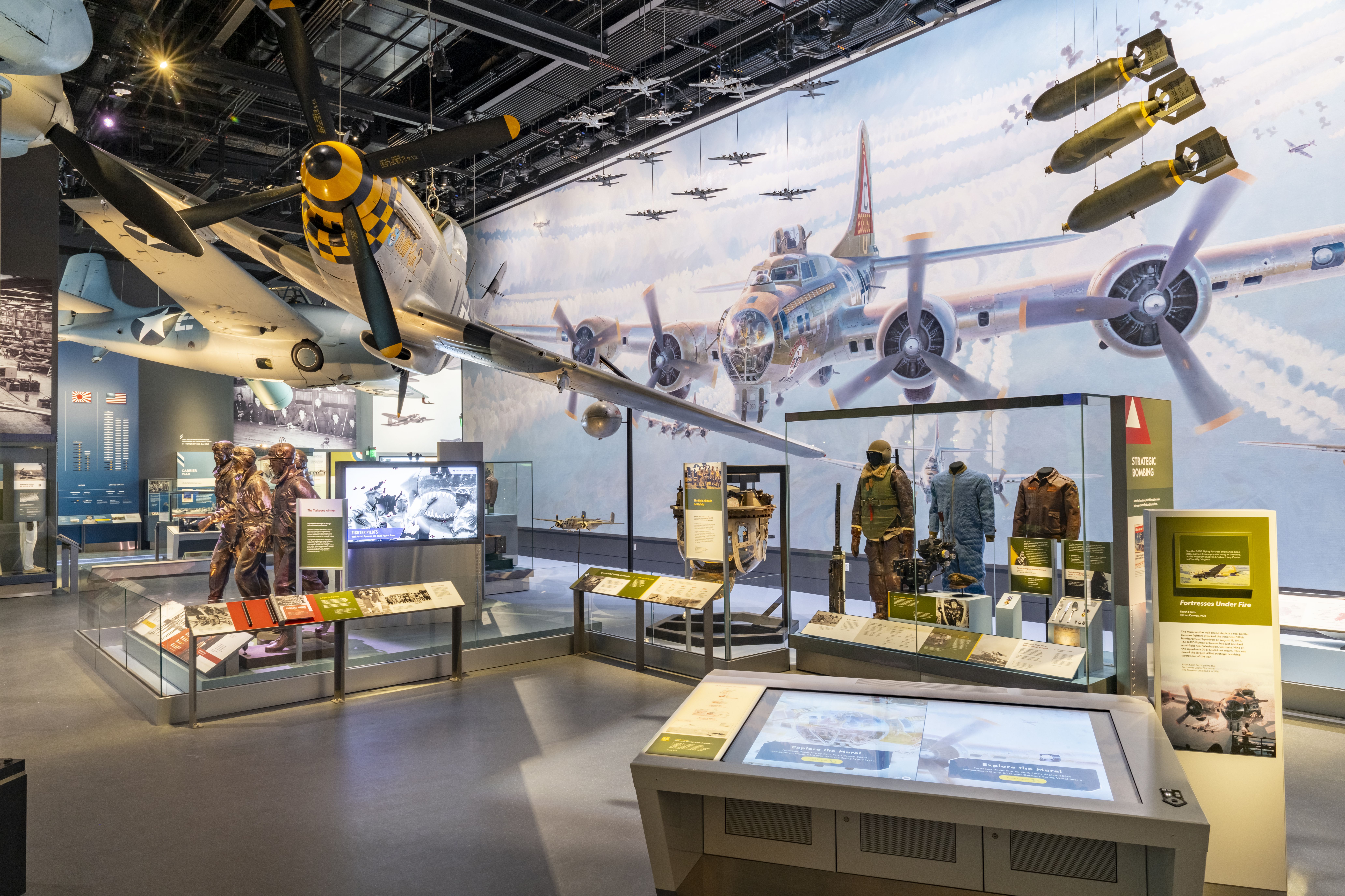 Museum exhibit showing a large WWII airliner mural with a propeller plane in the foreground, dozens of small planes suspended from the ceiling, hanging bombs on the right, and glass display cases with mannequins and info panels.