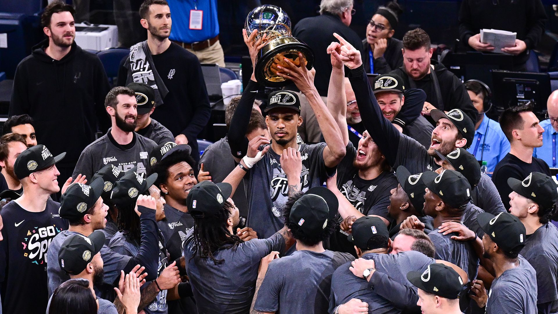 Spurs players lift the Western Conference finals trophy overhead, celebrating with teammates in gray shirts and caps.