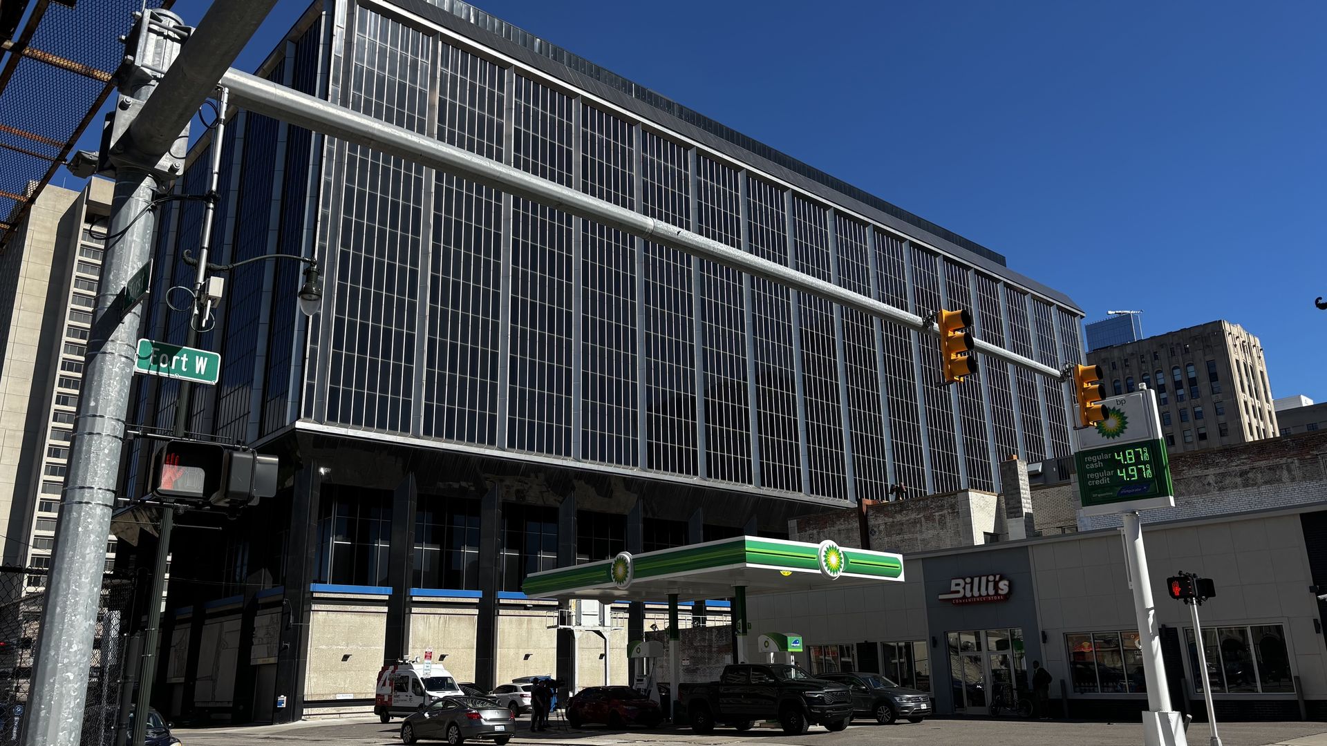 Tall glass office building with a grid of windows looms over a street intersection; in the foreground are a BP gas station canopy, a green "Fort W" street sign, and orange traffic lights under a clear blue sky.