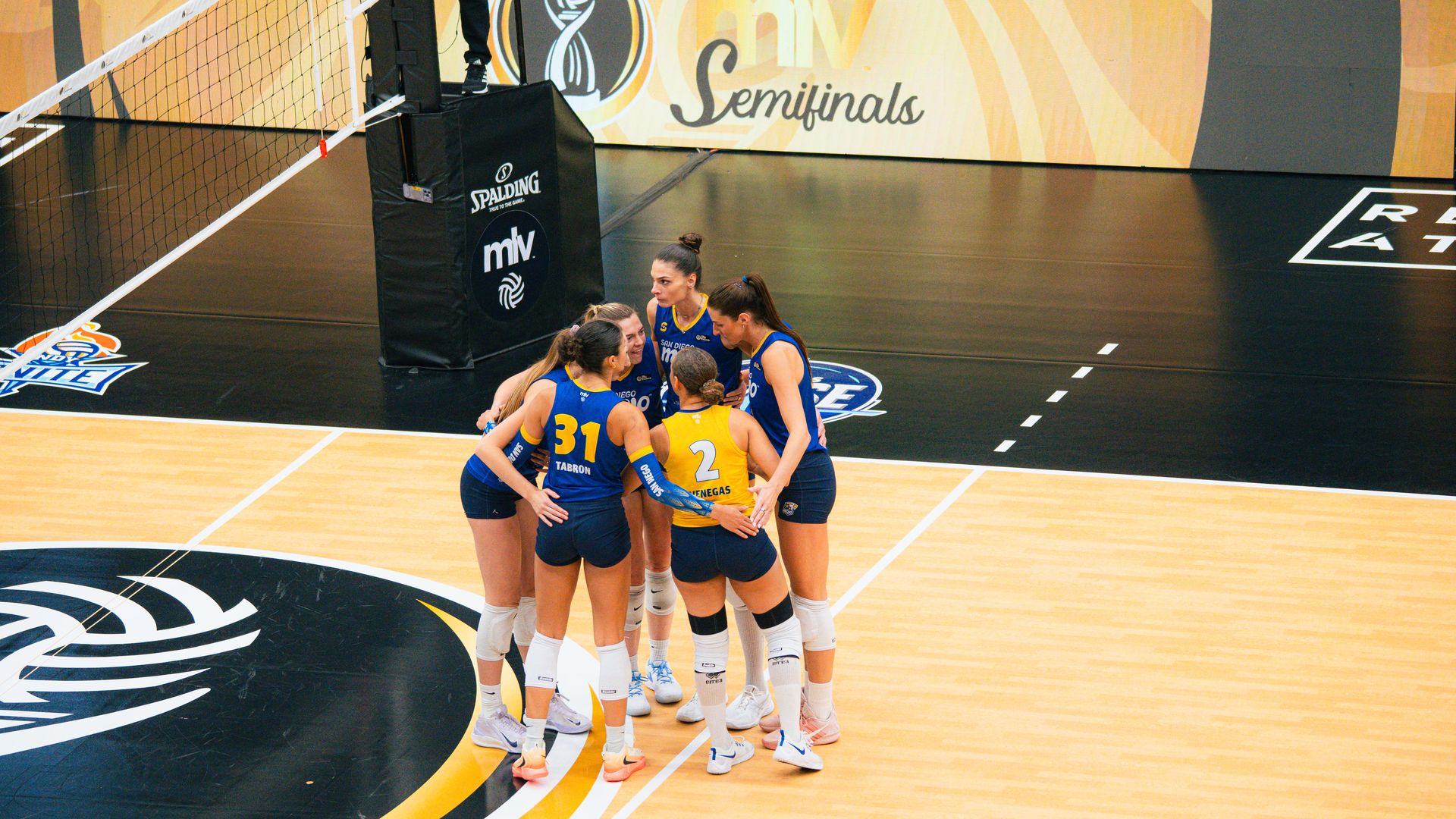 Group of women's volleyball players in blue and yellow uniforms huddle on a wooden court beside the net during a semifinal. A red digital timer shows 20:12 and a large banner reads Semifinals.