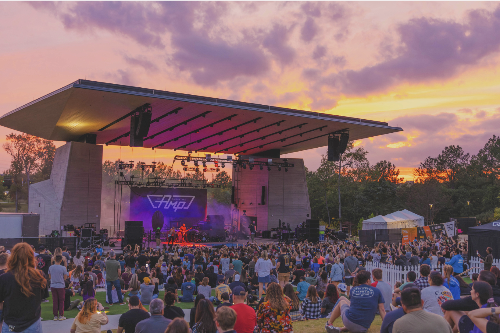 Outdoor concert at sunset with colorful sky, crowd watching band on stage under large roof, purple and orange lighting, trees and tents in background