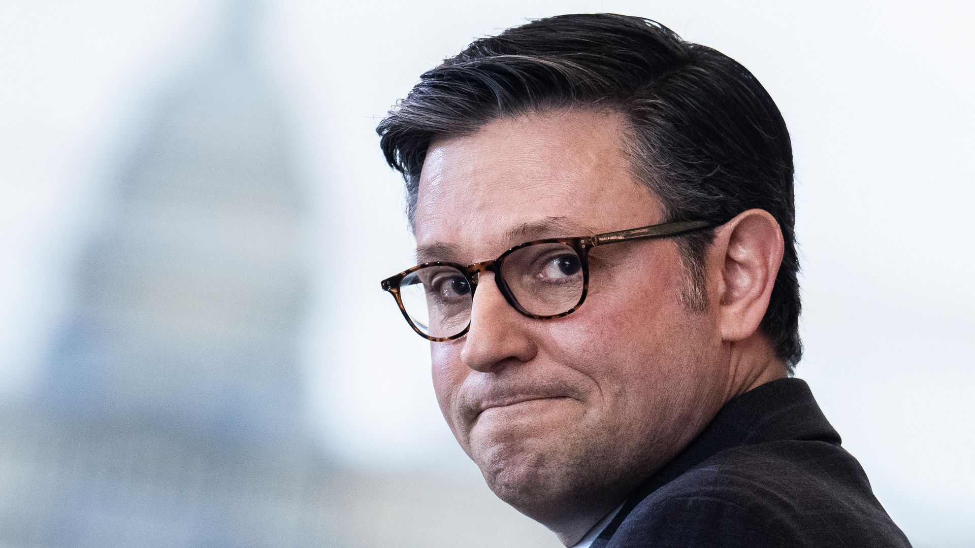 Speaker Mike Johnson, wearing a dark gray suit and glasses, with the Capitol building in the background.