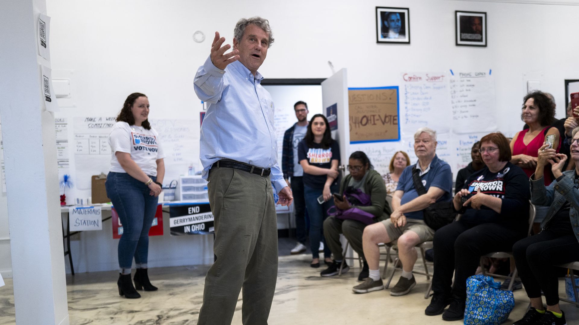 Man in light blue shirt and khaki pants speaking to seated and standing audience in a room with white walls covered in posters about voting and ending gerrymandering in Ohio.