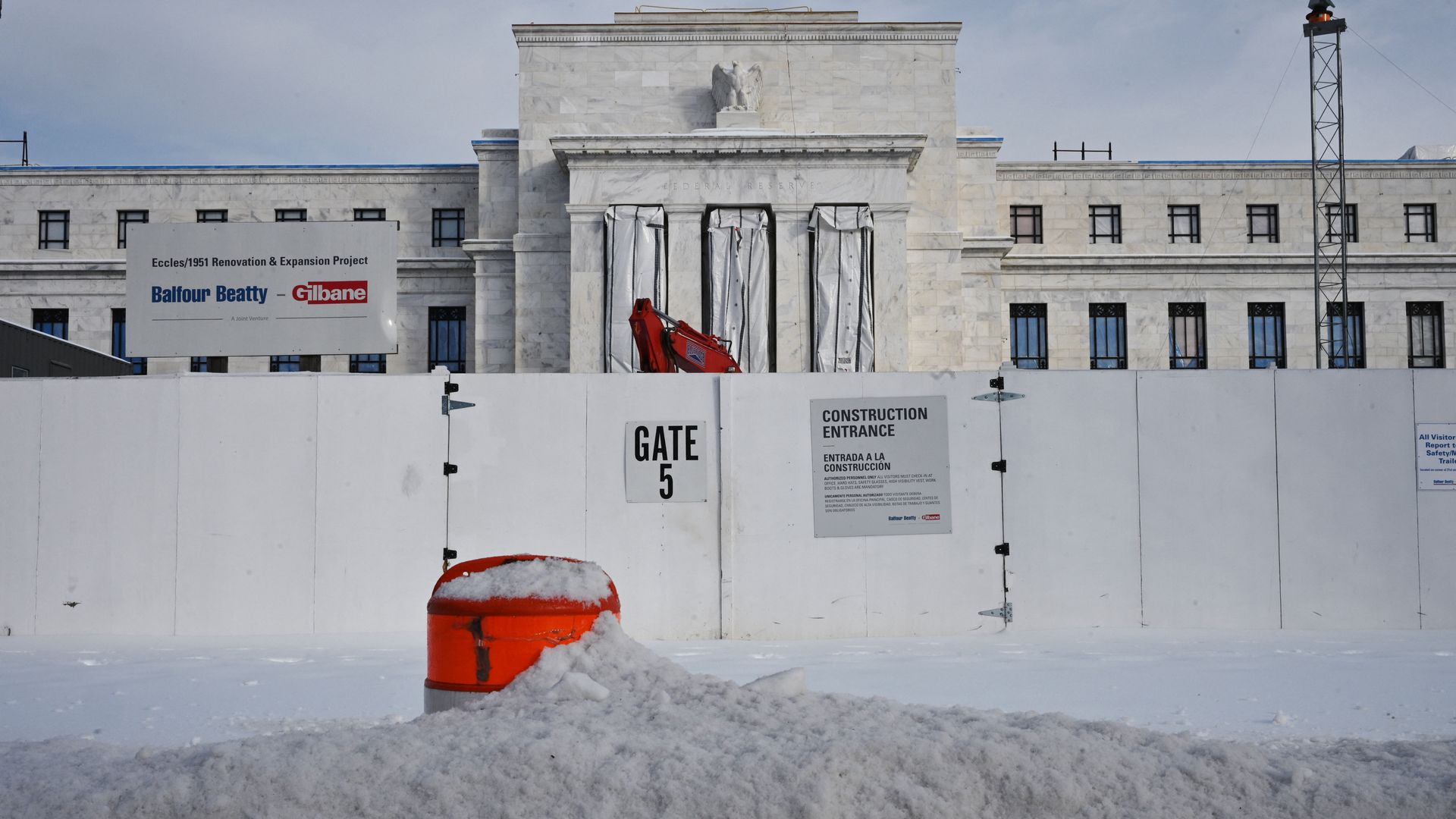 Snow-covered orange construction barrel in front of a white fence with signs reading "Gate 5" and "Construction Entrance" blocking a marble building under renovation.