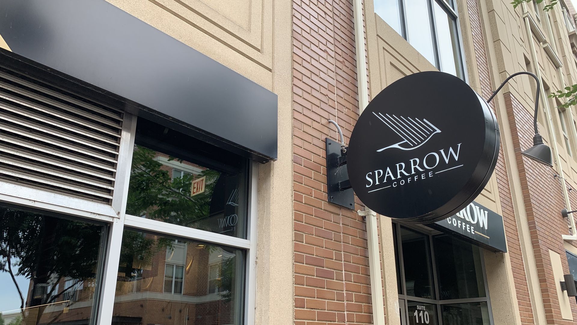 Round black Sparrow Coffee sign with a white sparrow motif and the words SPARROW COFFEE, mounted on a brick storefront beside large windows and a black awning.