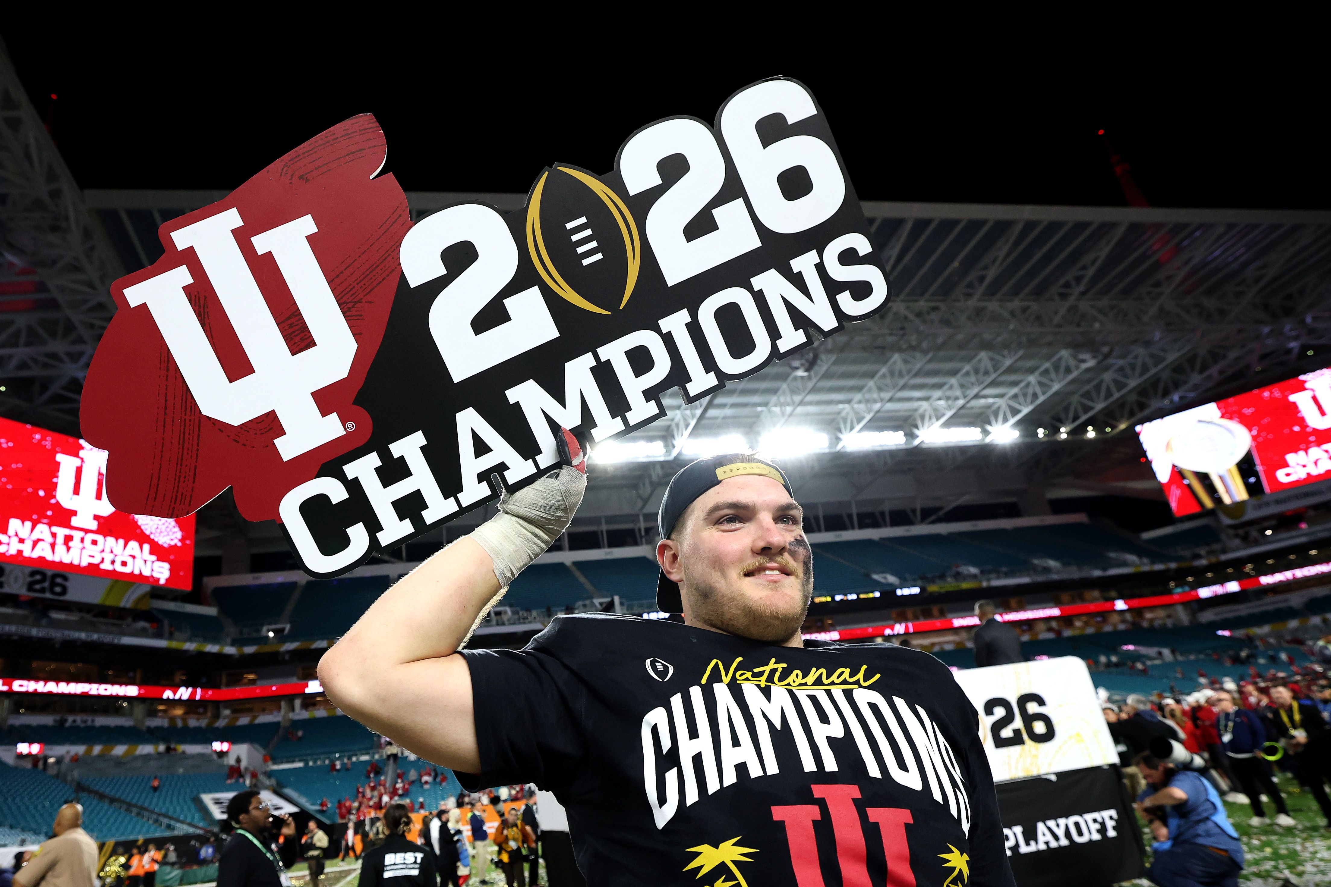 Man wearing black National Champions shirt holds large Indiana University 2026 Champions sign inside stadium at night with crowd in background.