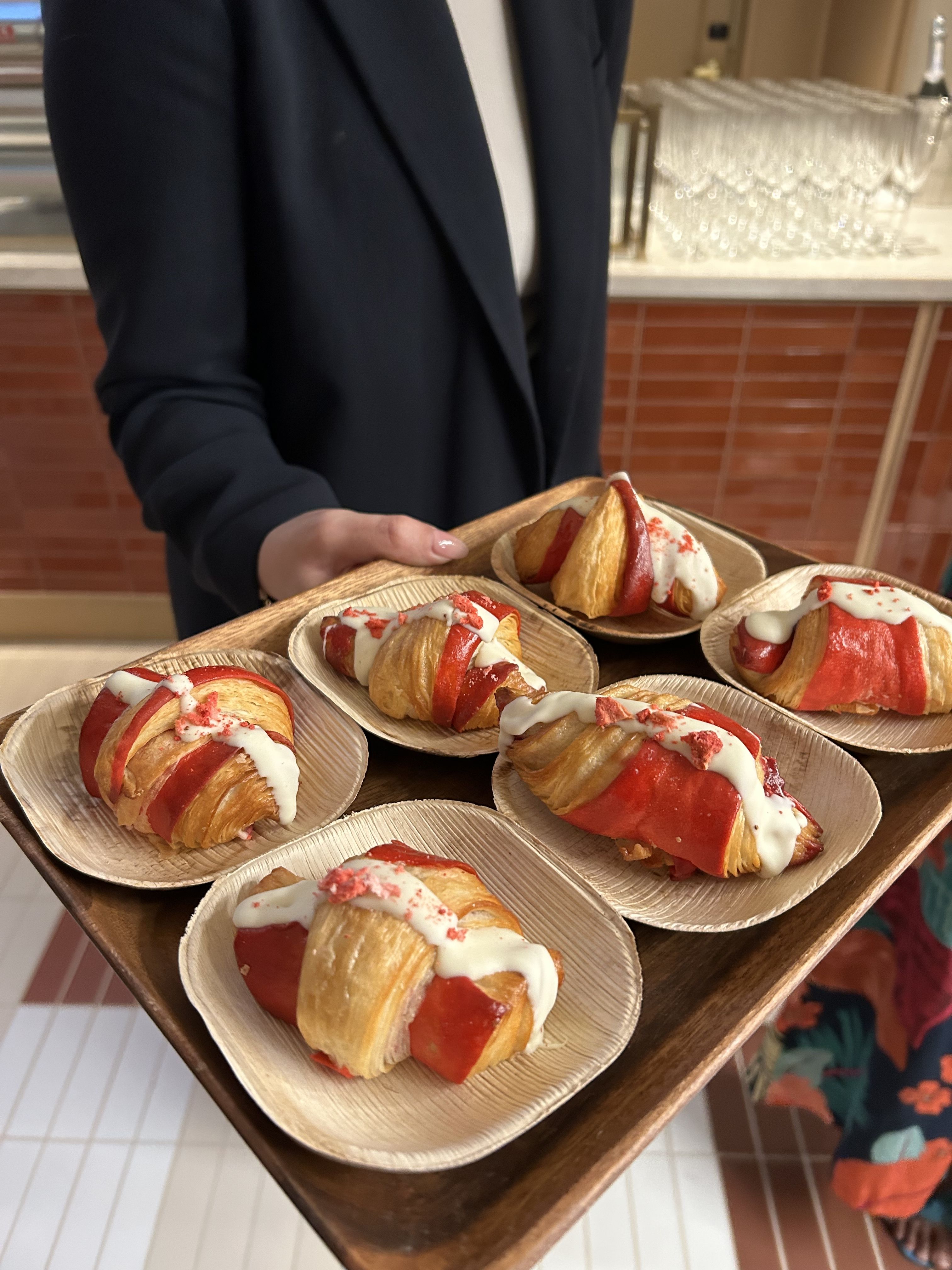 Person in a dark blazer holds trays of croissants glazed in red and topped with white icing; a row of glassware sits on a counter in the background.