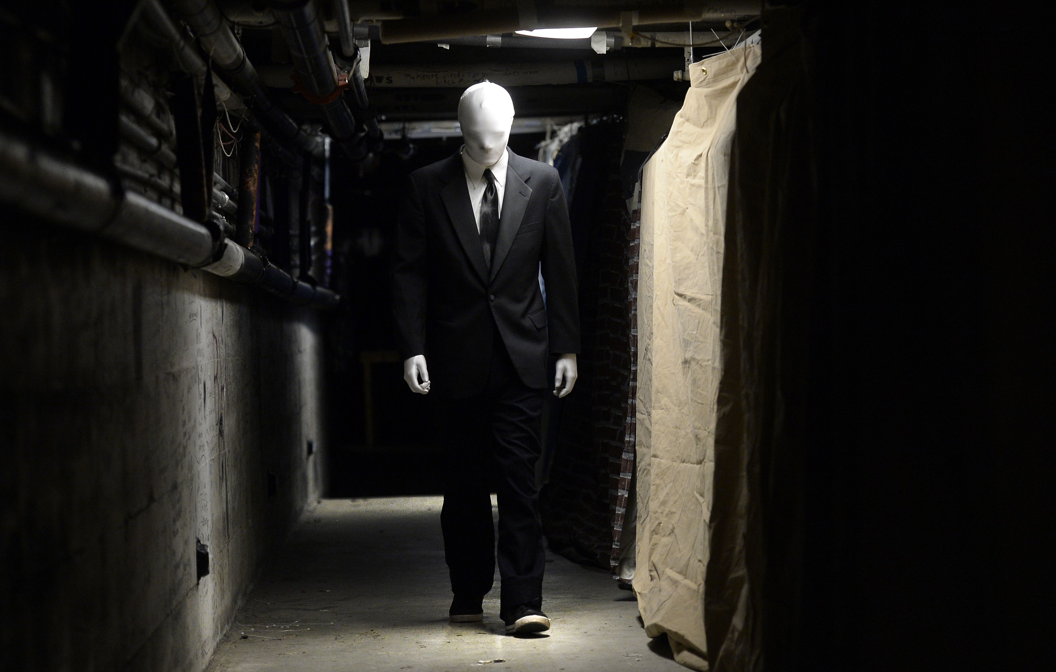Figure with a white featureless head wearing a black suit and tie walking down a dimly lit, narrow industrial hallway with pipes and draped fabric on the walls.