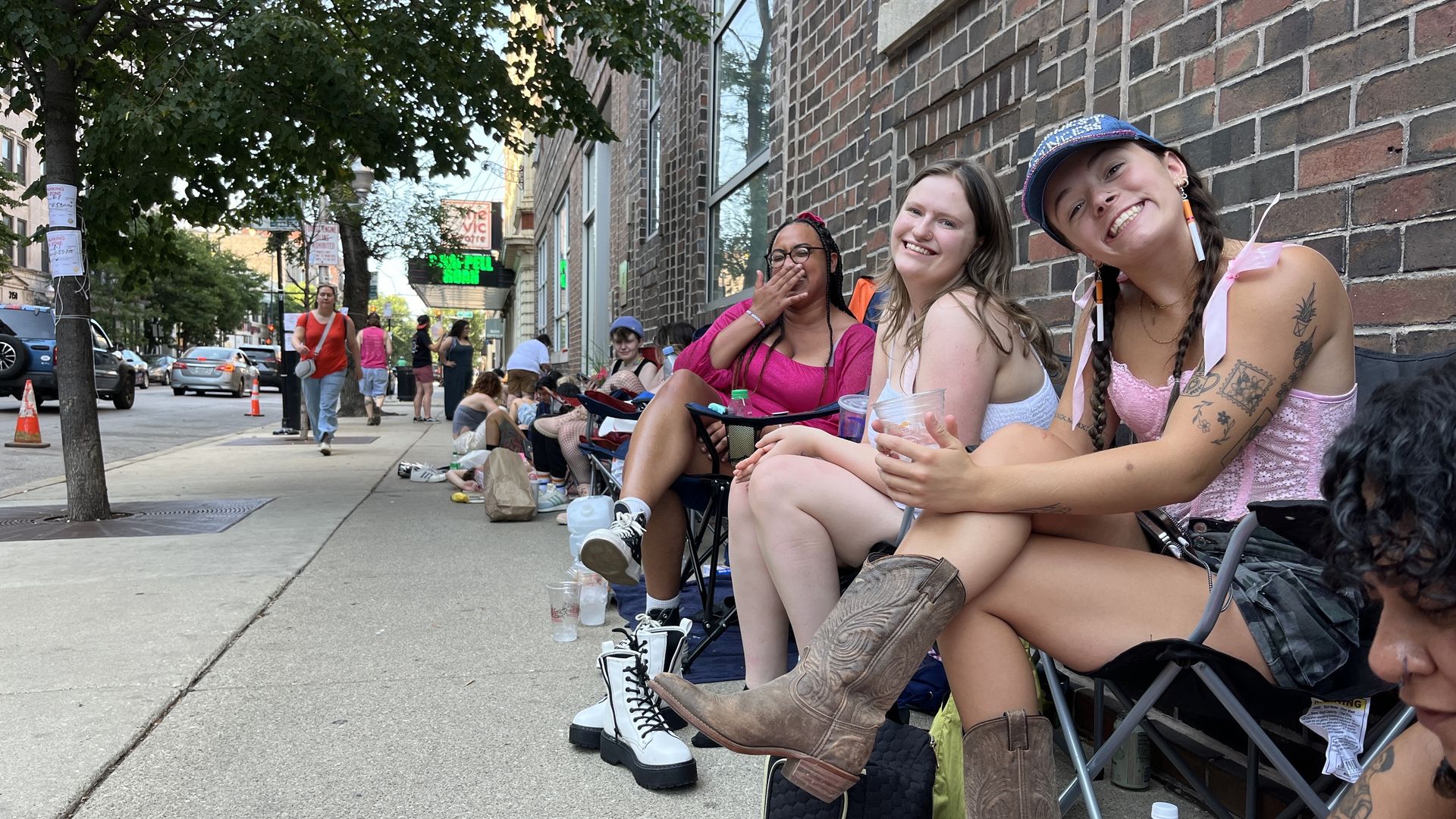 Photo of people sitting in lawn chairs in line outside a building 