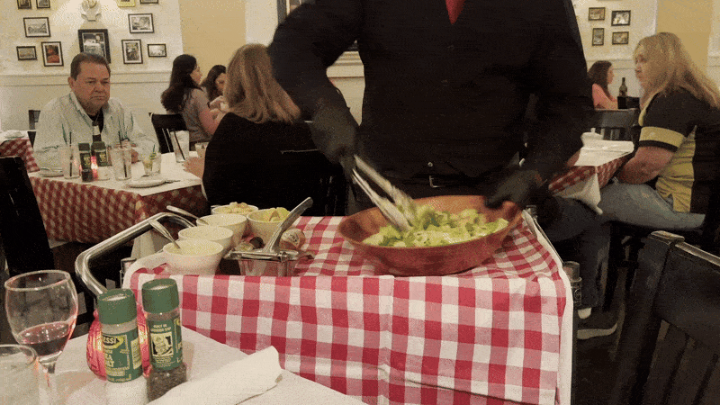 A person makes a Caesar salad at a wheeled-in cart next to a dining table.