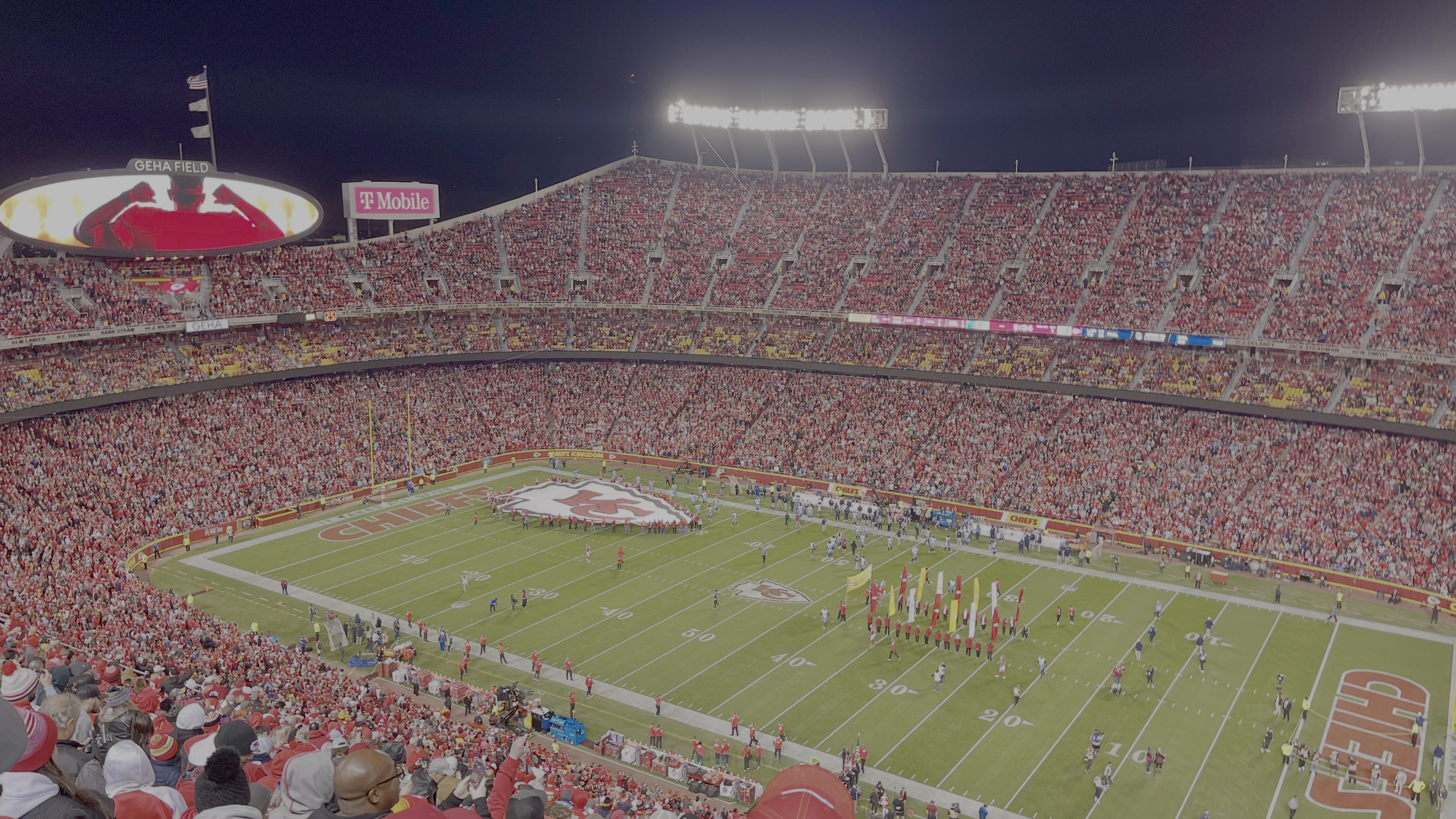 Nighttime stadium packed with red-clad fans, bright floodlights, and a large oval screen displaying "GEHA FIELD"; players on the green field and a marching band along the sideline.