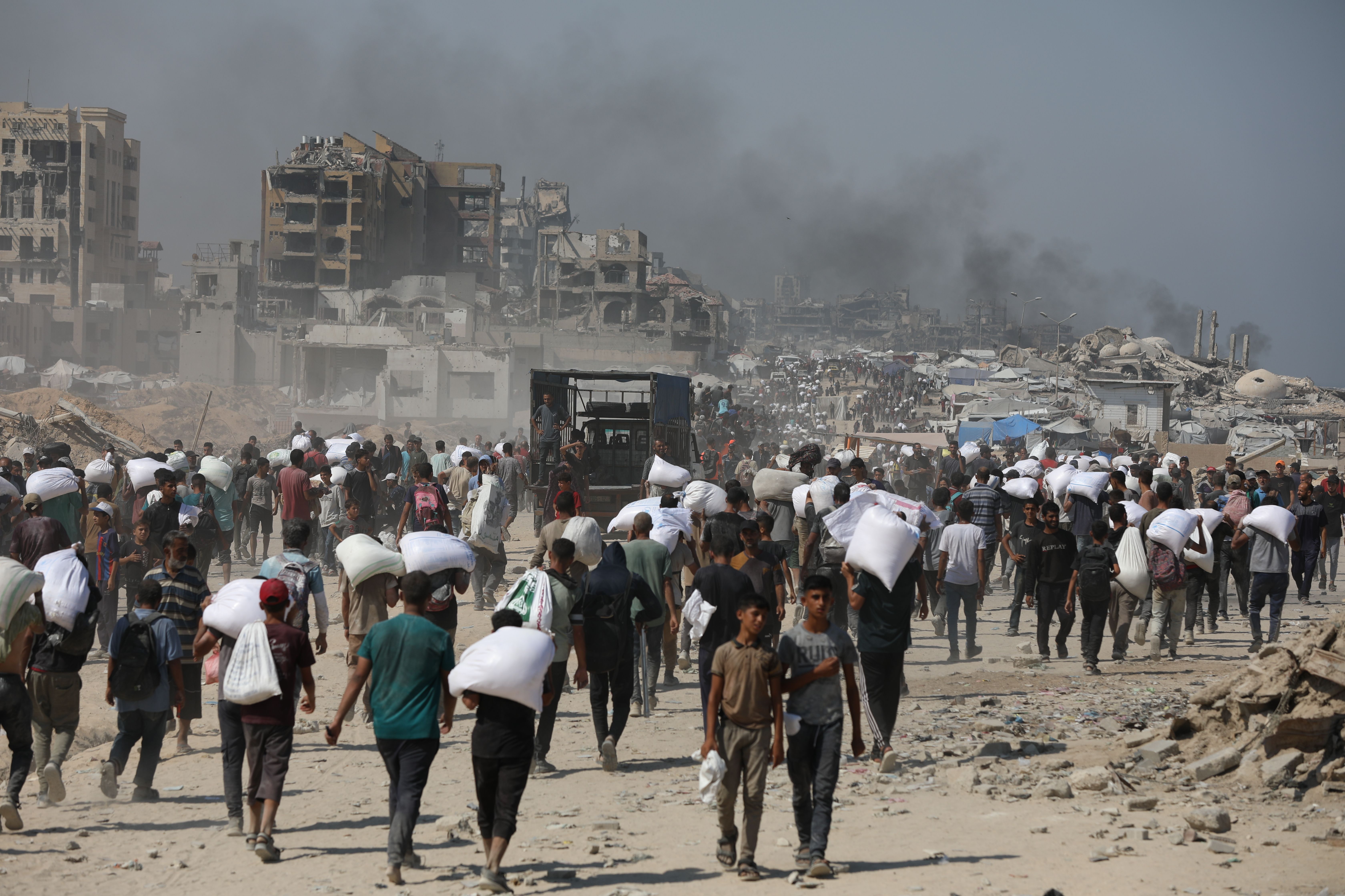Crowd of people carrying white bags walking through a dusty street with heavily damaged buildings and smoke in the background under a clear blue sky.