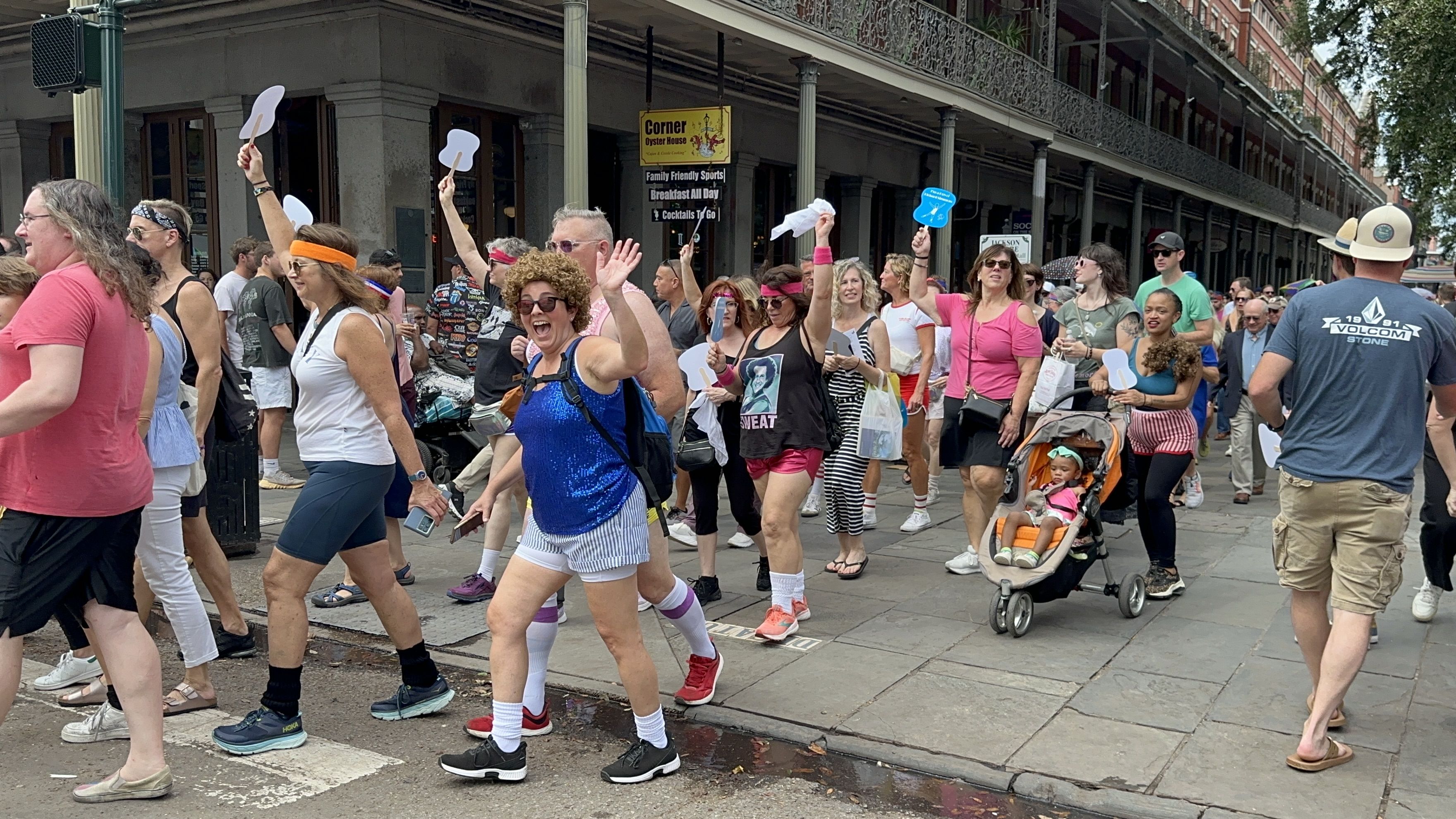 People wave to the camera as they walk past. Some are dressed in 1980s-style workout gear and wigs.