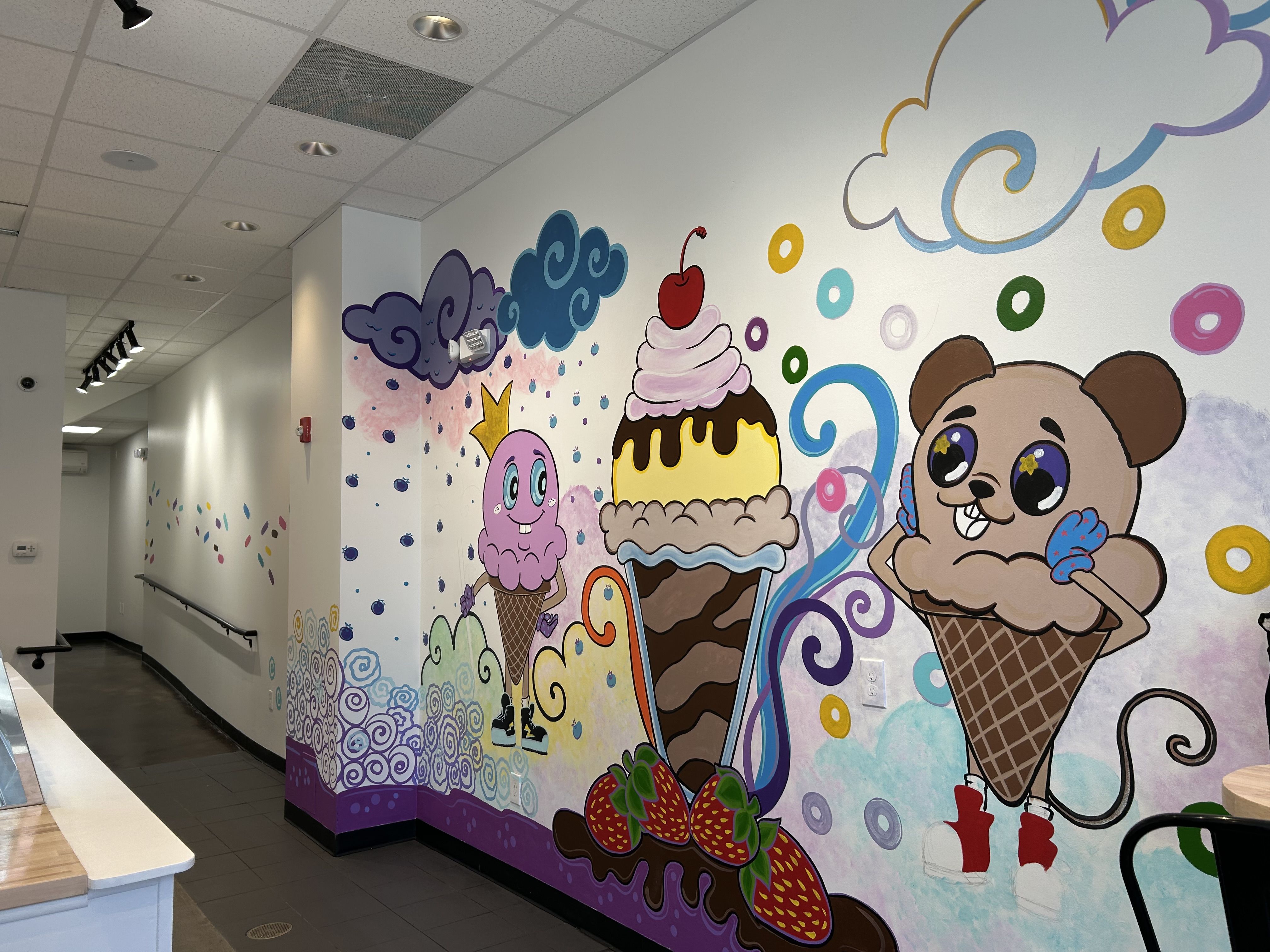 Colorful mural in an ice cream shop featuring cartoon ice cream cones with faces, a mouse character, large strawberries, swirls, clouds, and bright circular patterns on white walls.