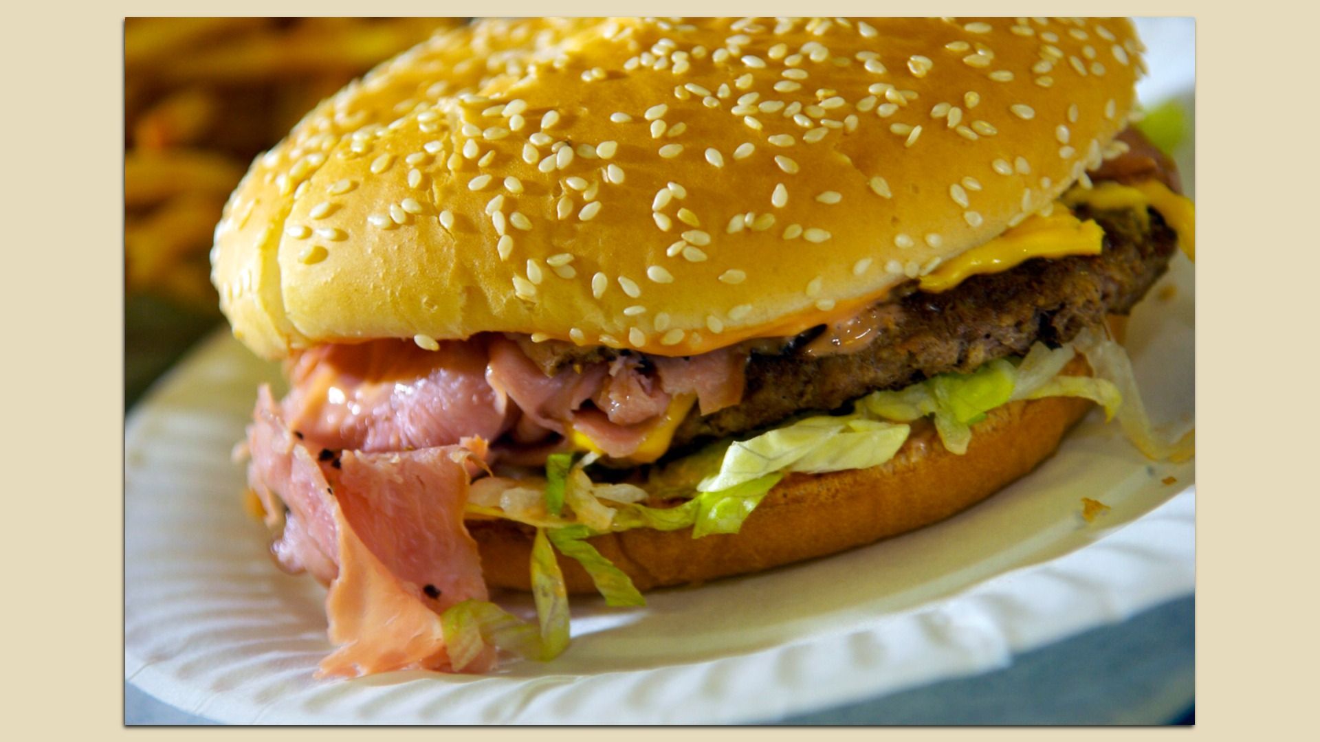 Close-up of a sesame-seed hamburger with melted cheese, a beef patty, lettuce, and pink ham peeking from a toasted bun on a white plate.