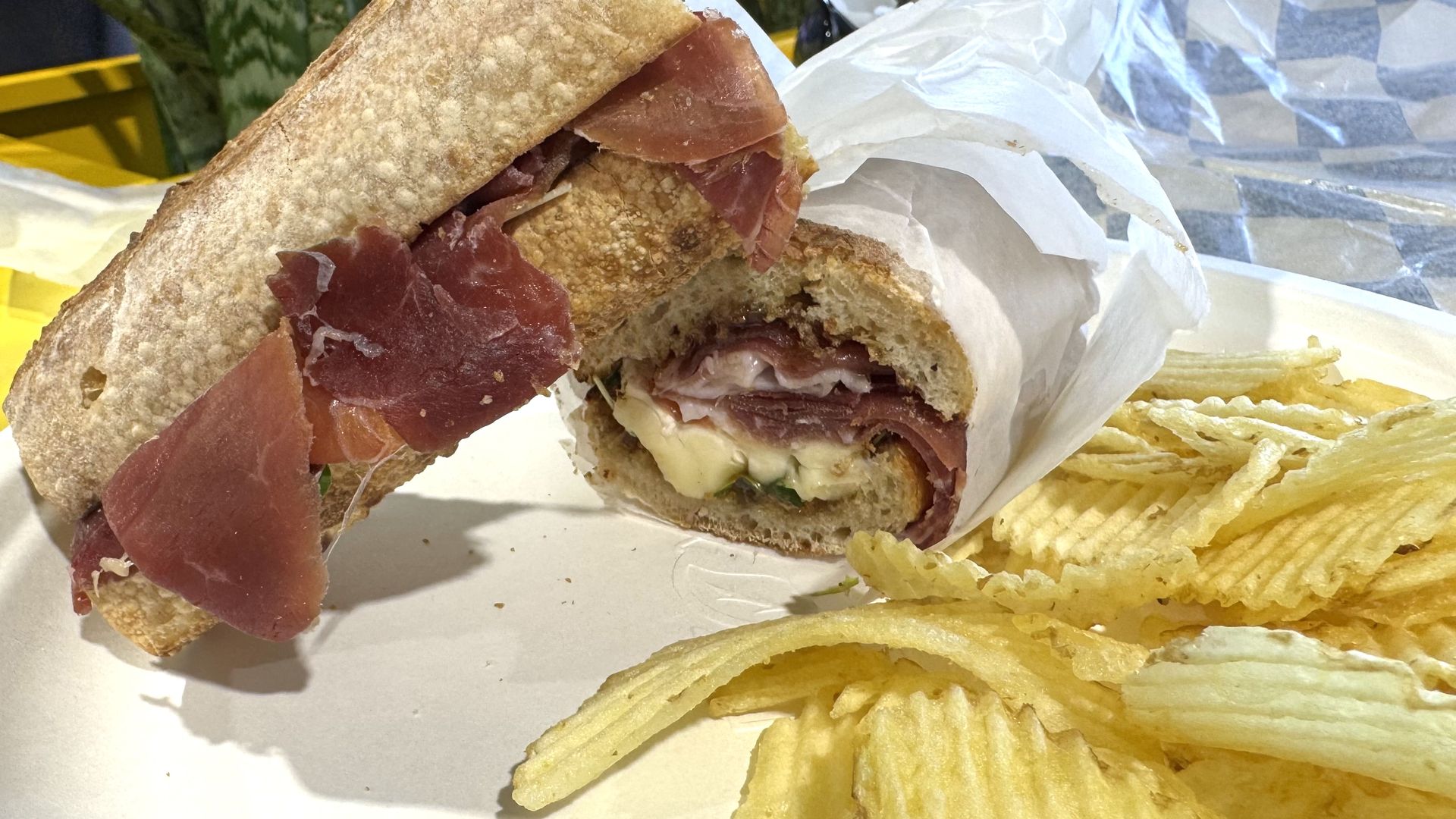 Cross-section of a crusty baguette sandwich with thin prosciutto ham and melted cheese, wrapped in white paper, served with crinkle-cut potato chips on the side.